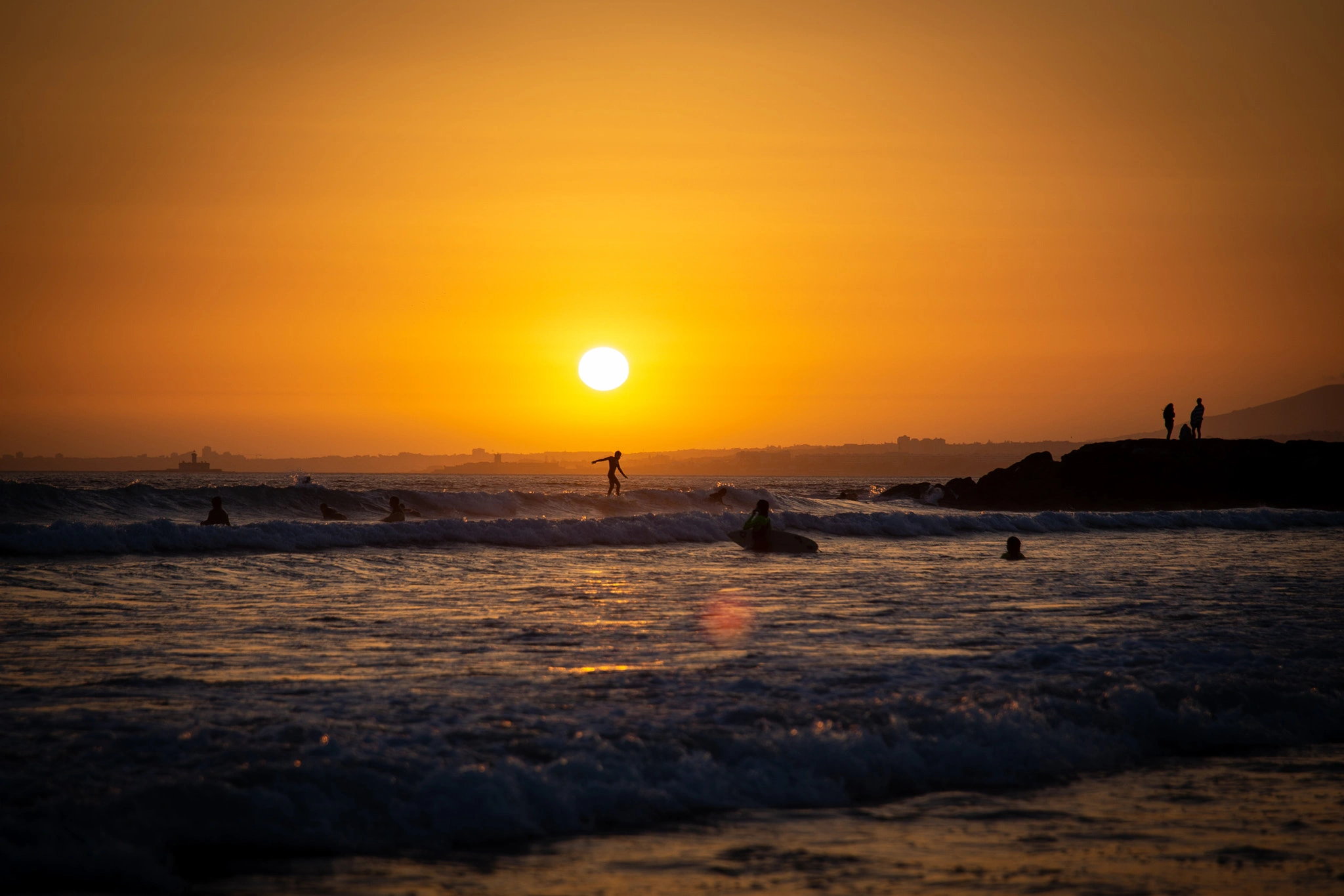 Several surfers scattered across the ocean as the sun sinks to the horizon, one standing on a small wave while silhouettes watch from the rocks — the whole scene bathed in deep amber.