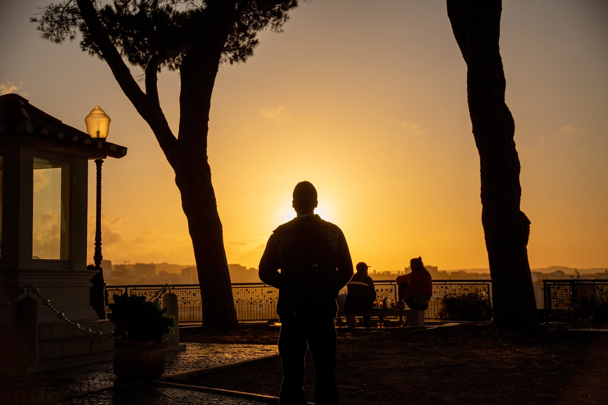 A lone figure with a backpack stands silhouetted at a Lisbon viewpoint, the city rooftops and river dissolving into a blazing golden haze behind ornate ironwork and stone pines.
