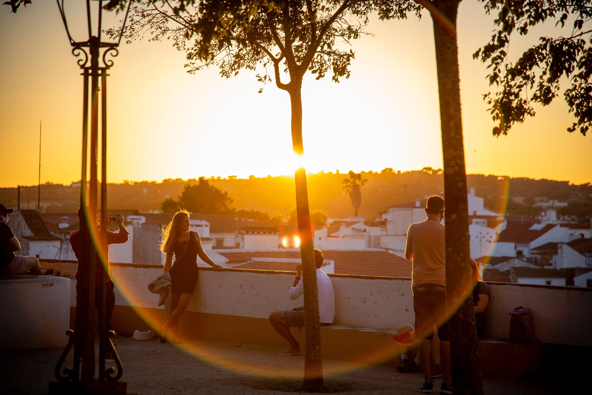 Young people gather at a hilltop viewpoint as golden light floods a white-walled Alentejo town below, a circular iron gate framing the scene and a blazing sun flaring into the lens.