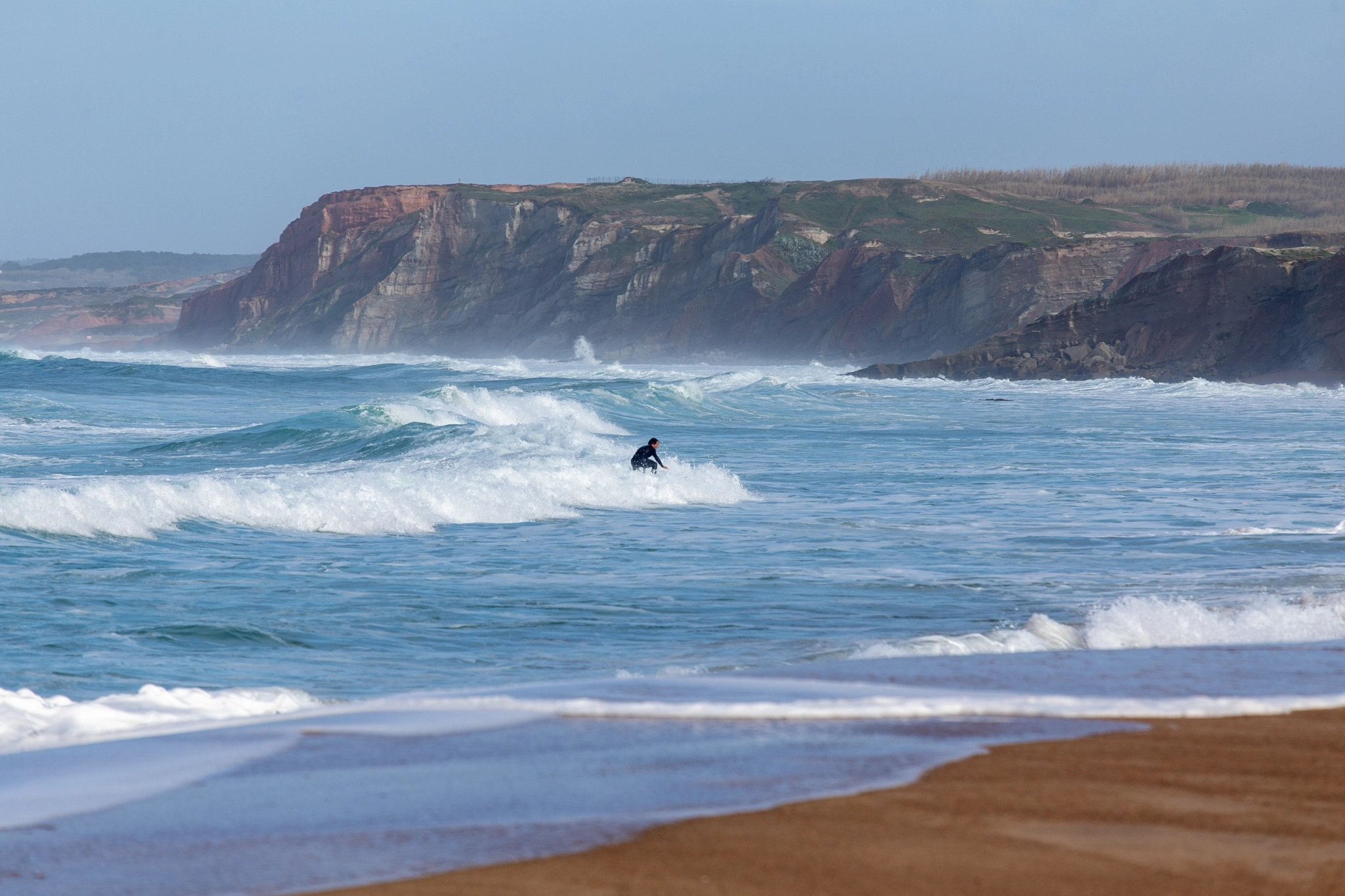 A lone surfer paddles hard to meet a breaking wave, the red sandstone cliffs of the Portuguese coast rising behind a wide arc of white water.