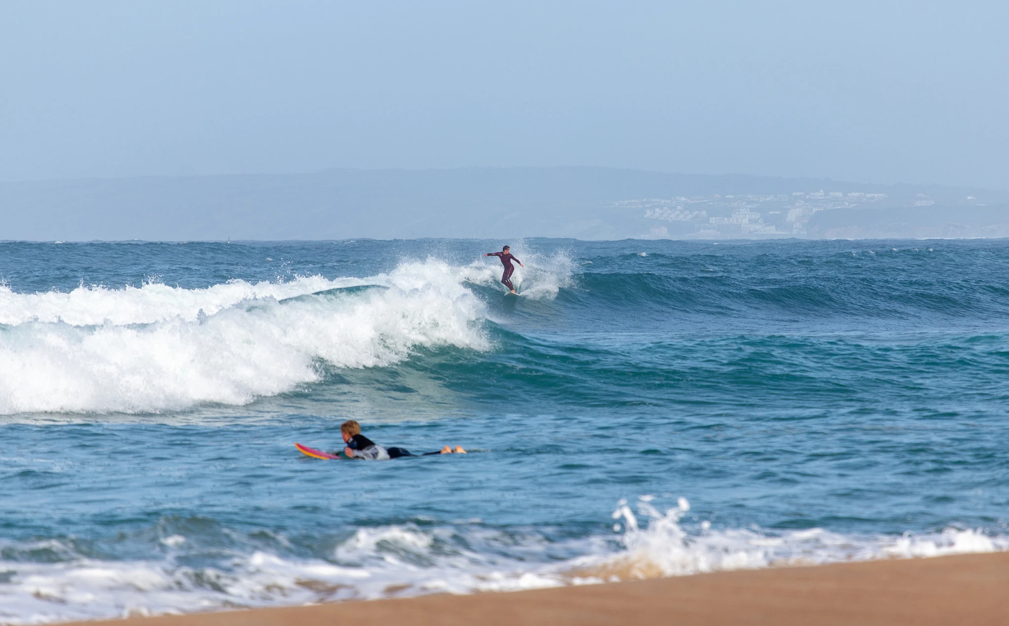 A surfer trims across a clean blue-green face while another paddles beneath in the foreground, a misty coastal town barely visible on the horizon.
