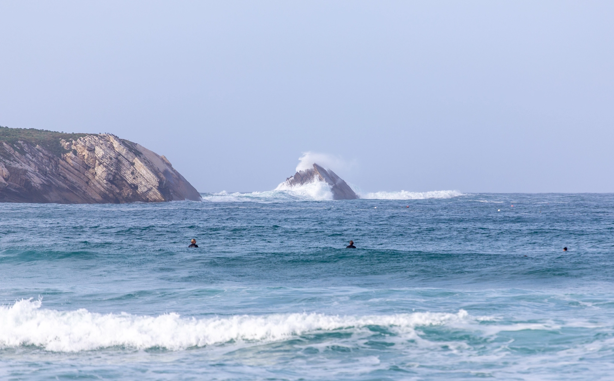 Three surfers wait in the lineup as a wave detonates against a solitary sea stack behind them, the red cliffs of the headland framing the whole chaotic scene.