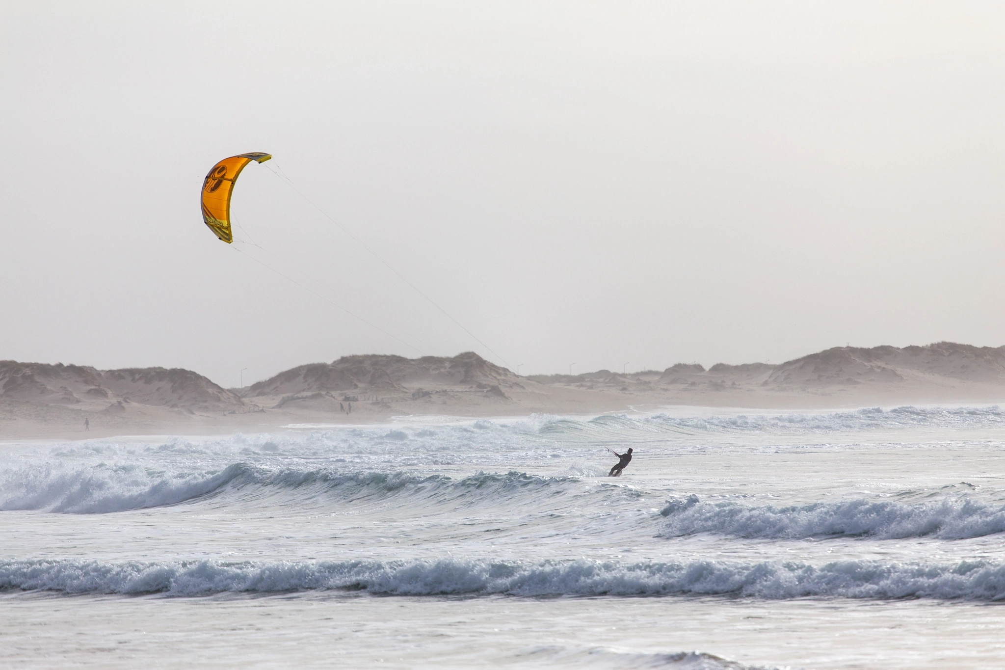 A kitesurfer with a vivid orange canopy carves through stormy grey chop, sand dunes stretching behind in a bleak, windswept landscape.