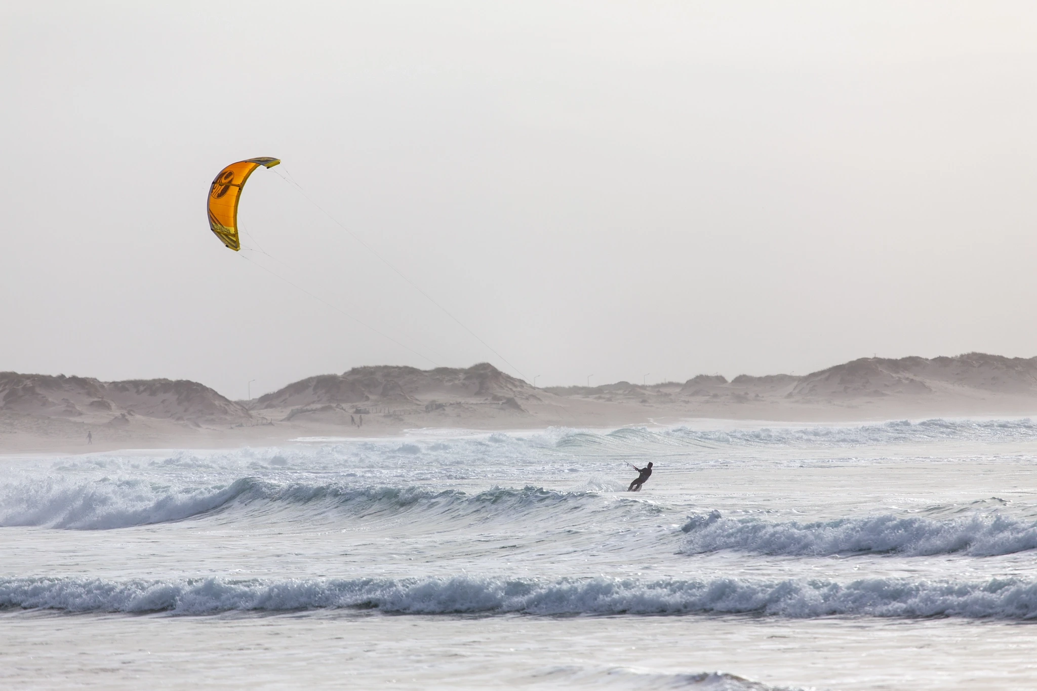 A kitesurfer with a vivid orange canopy carves through stormy grey chop, sand dunes stretching behind in a bleak, windswept landscape.