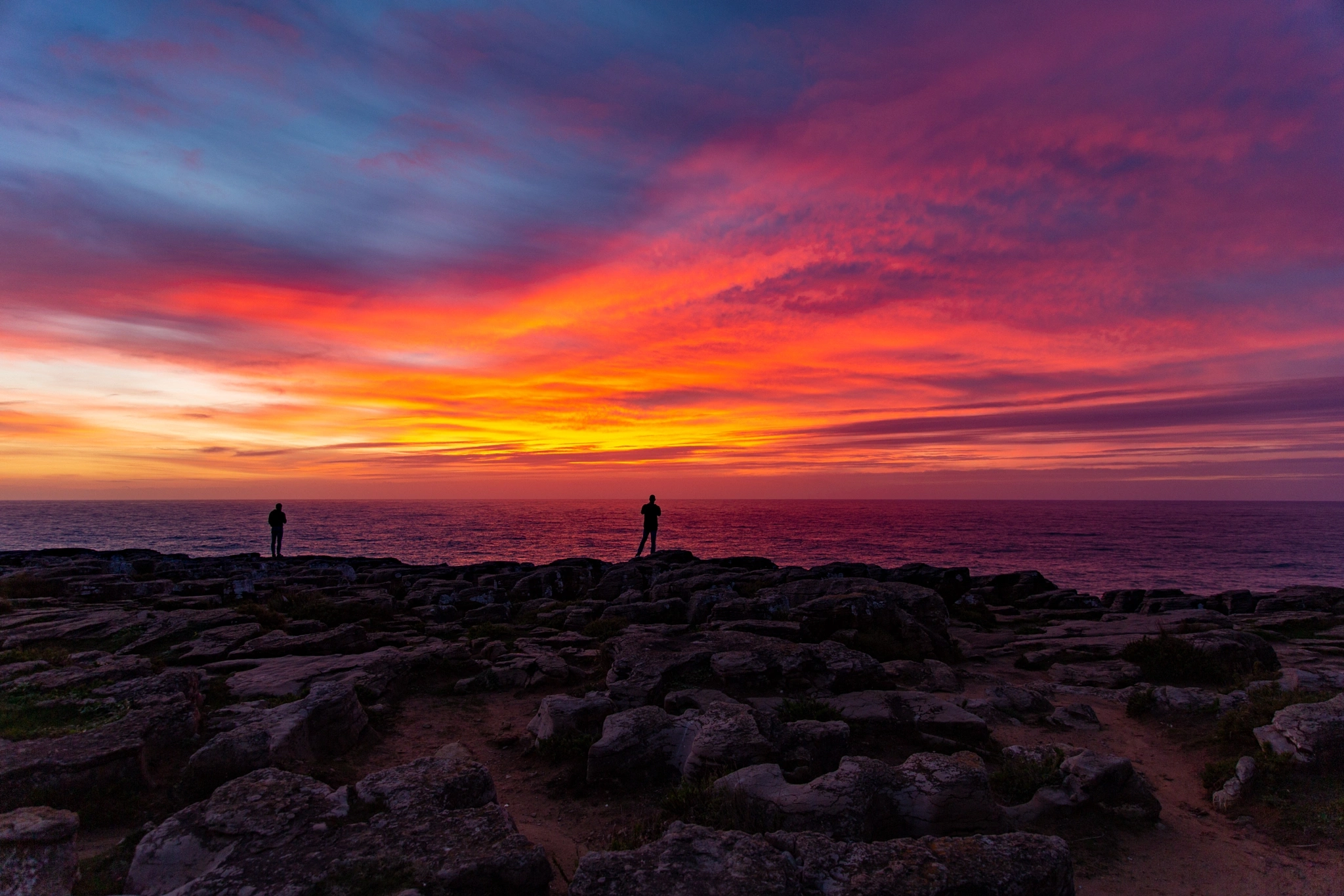 Two figures stand motionless on a fractured coastal plateau as the sky ignites in violet, crimson and gold behind them, the Atlantic horizon ablaze with the last light of day.