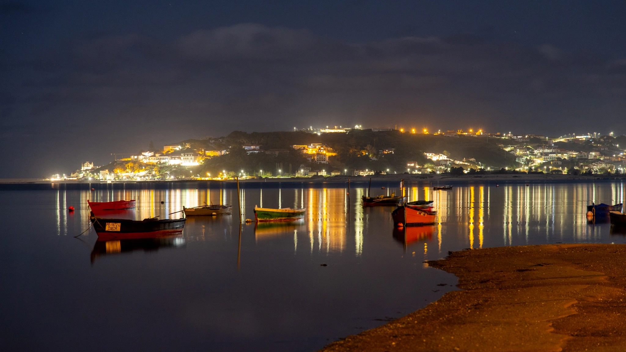 Painted fishing boats rest on the glassy surface of a harbor at night, the town's lights bleeding into long golden reflections across the still water.