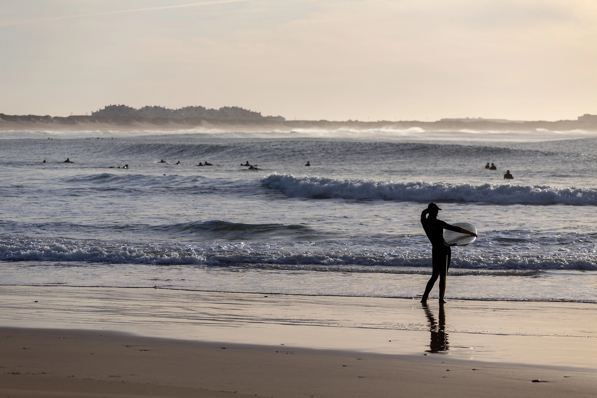 A surfer stands at the shoreline studying the break, board at their side, while a line of other surfers waits in the distant haze — a quiet moment of assessment before paddling out.