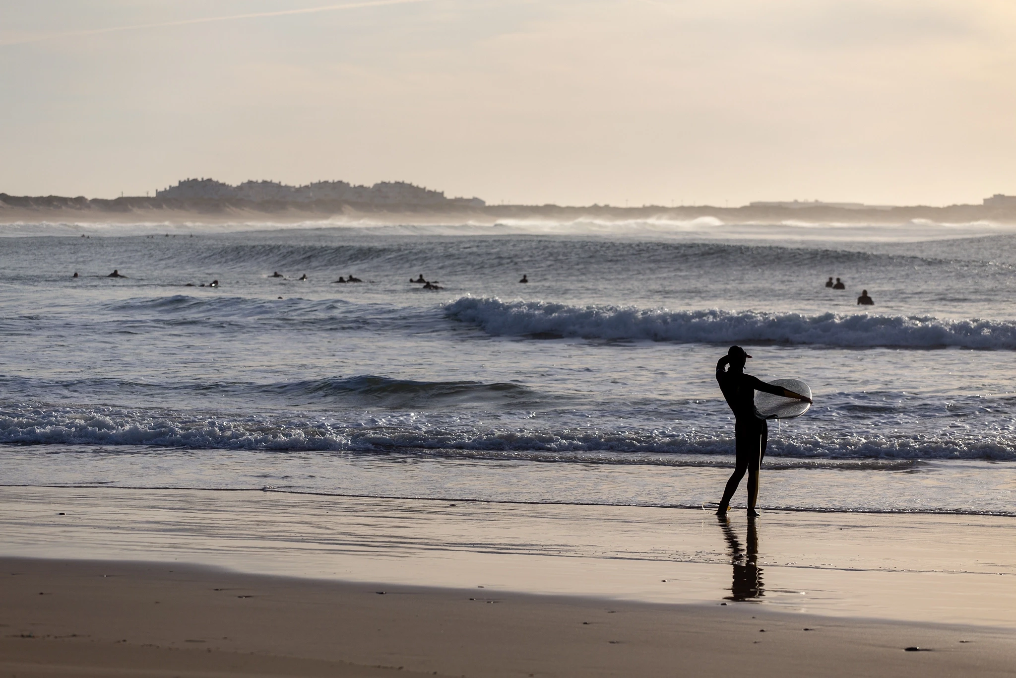 A surfer stands at the shoreline studying the break, board at their side, while a line of other surfers waits in the distant haze — a quiet moment of assessment before paddling out.