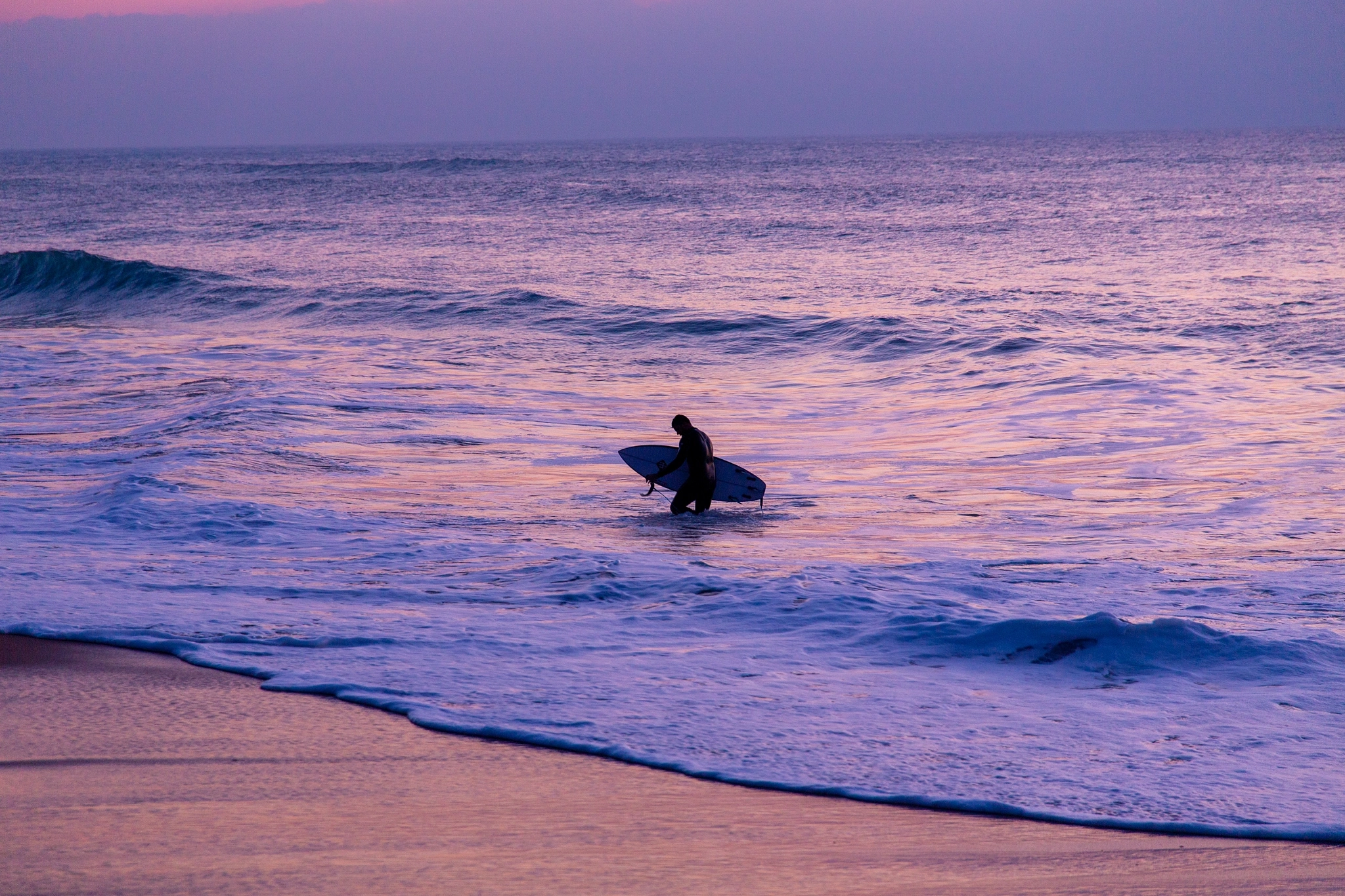 A surfer wades back through the shallows at dusk, the whole ocean suffused in deep violet and rose, the last light painting the water and wet sand in cool, melancholic tones.