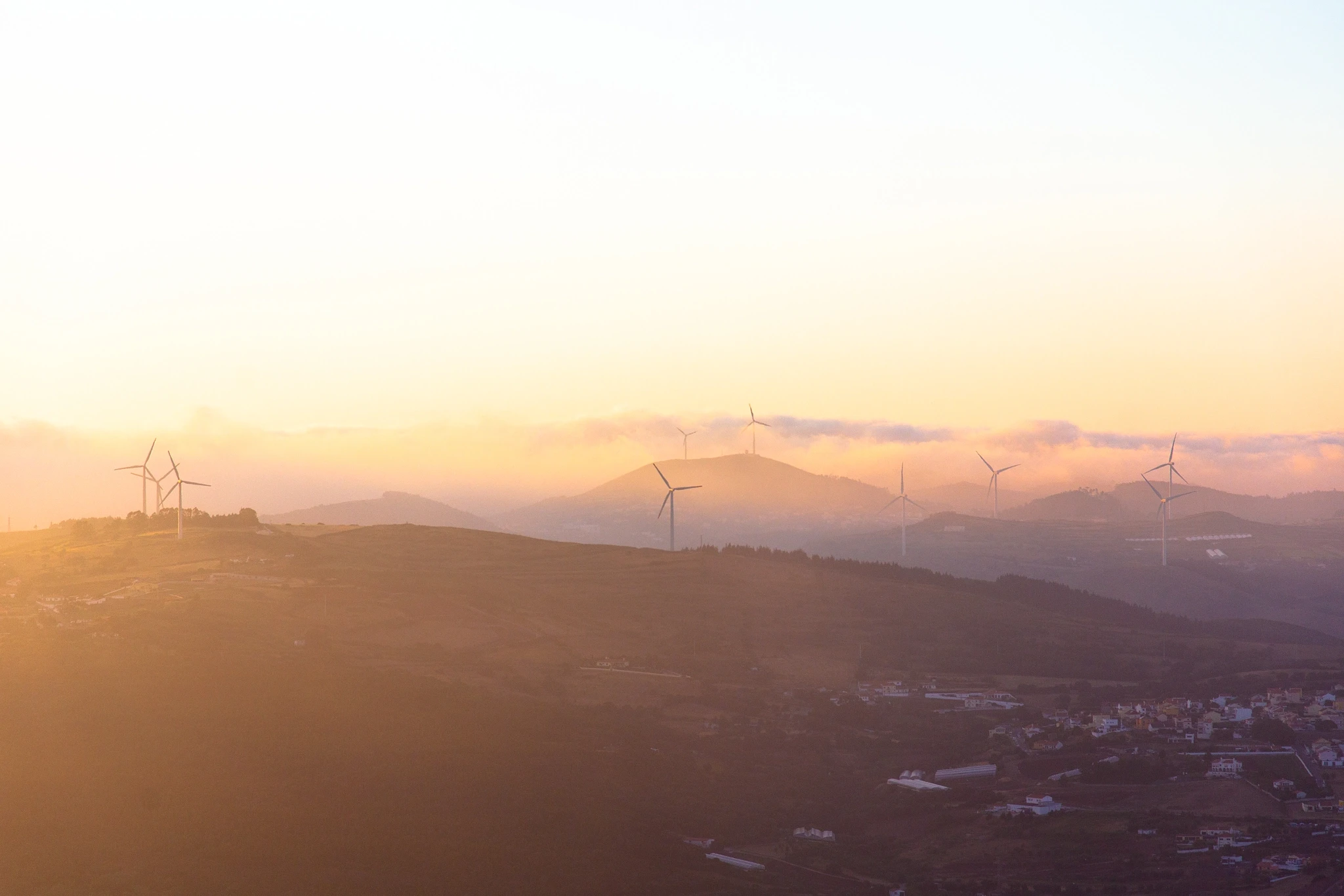 Wind turbines emerge from a warm misty haze over layered mountain ridges at sunrise, their white blades catching the first gold of the day against fading hillside silhouettes.