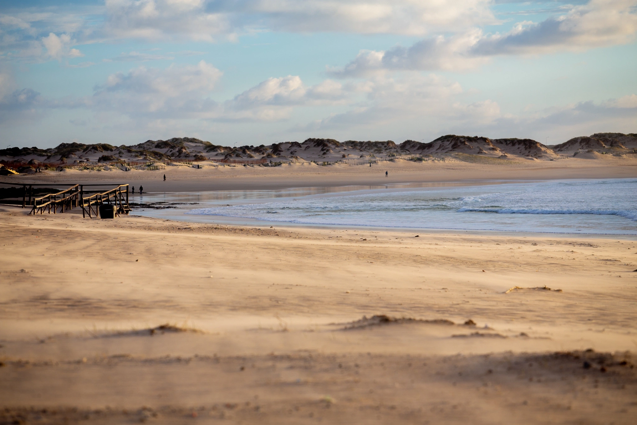 A wooden boardwalk cuts through windswept grass toward a vast, deserted beach, sandy dunes rolling away to the horizon under a dramatic cloudy sky.