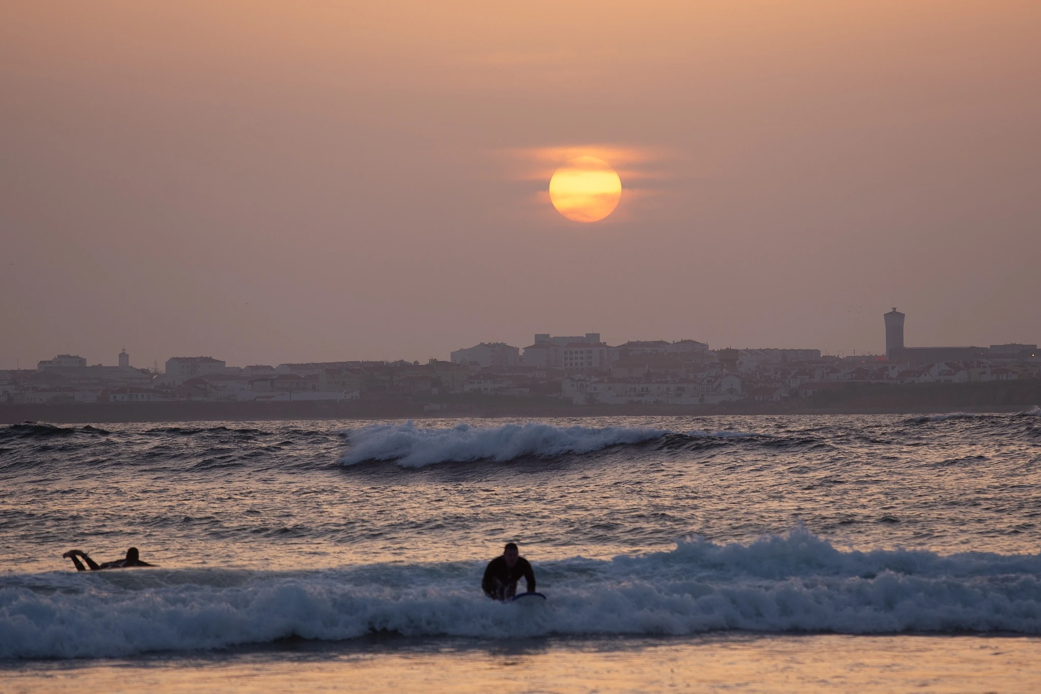 Two surfers float in the lineup as the sun sets behind a hazy city skyline, the orange globe reflected in the flat water between breaking waves.