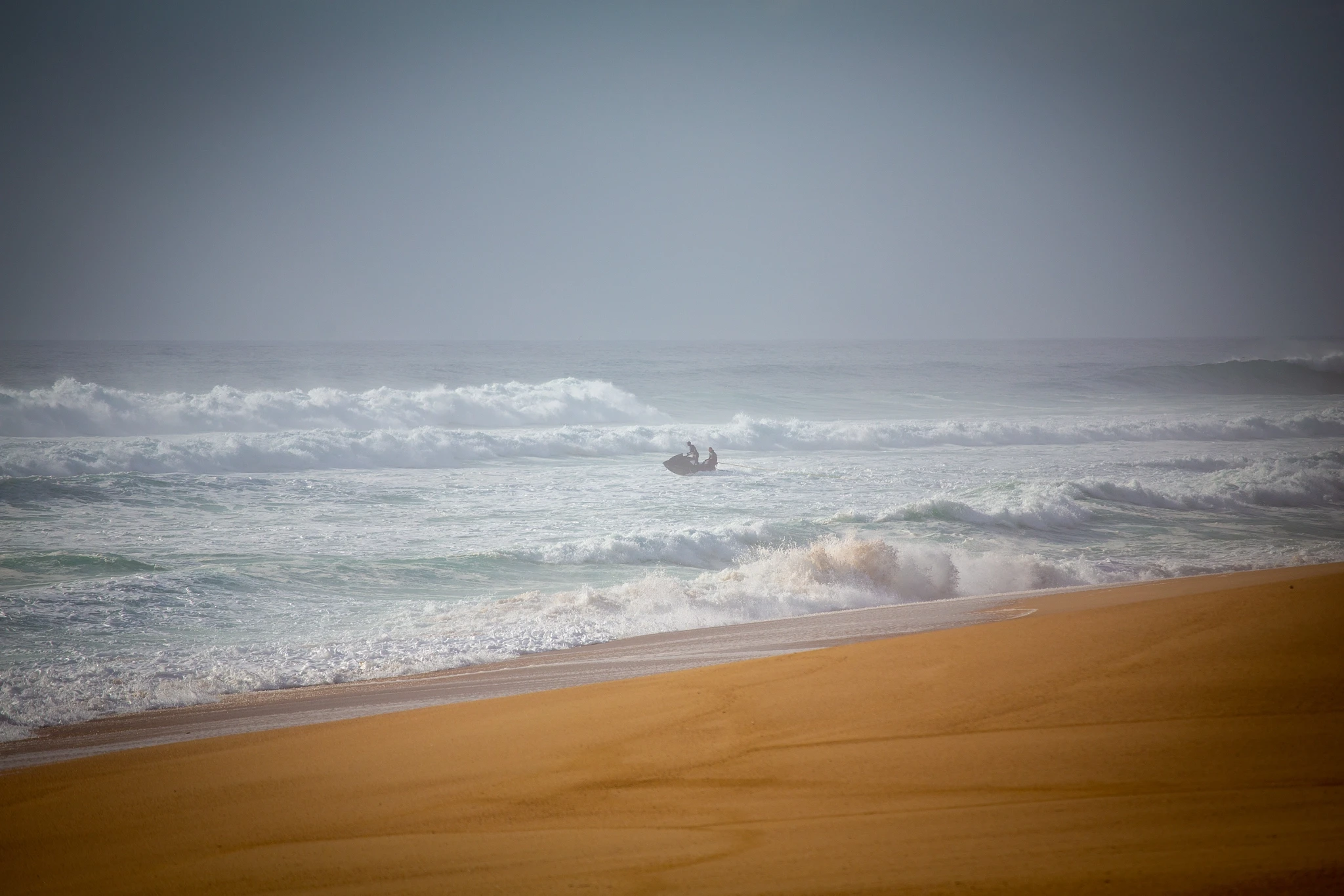A tow-in jet ski disappears into a wall of churning white water under a pewter sky, the scale of the swell reducing it to a tiny speck against the chaos of the Atlantic.