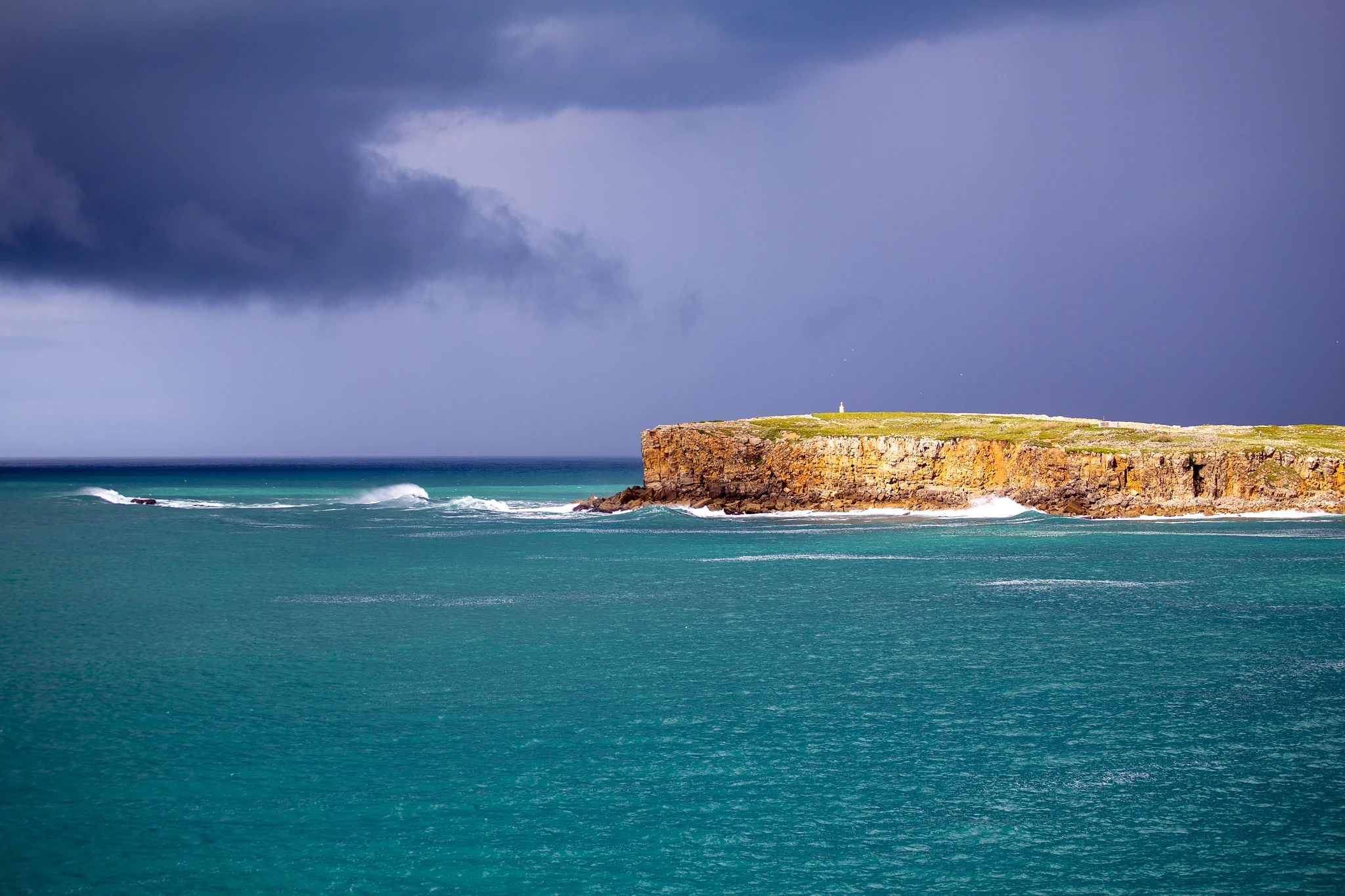 A table-flat island with a solitary lighthouse glows against a seething indigo storm front, the sea between glowing an improbable turquoise — calm water and wild sky in violent contrast.