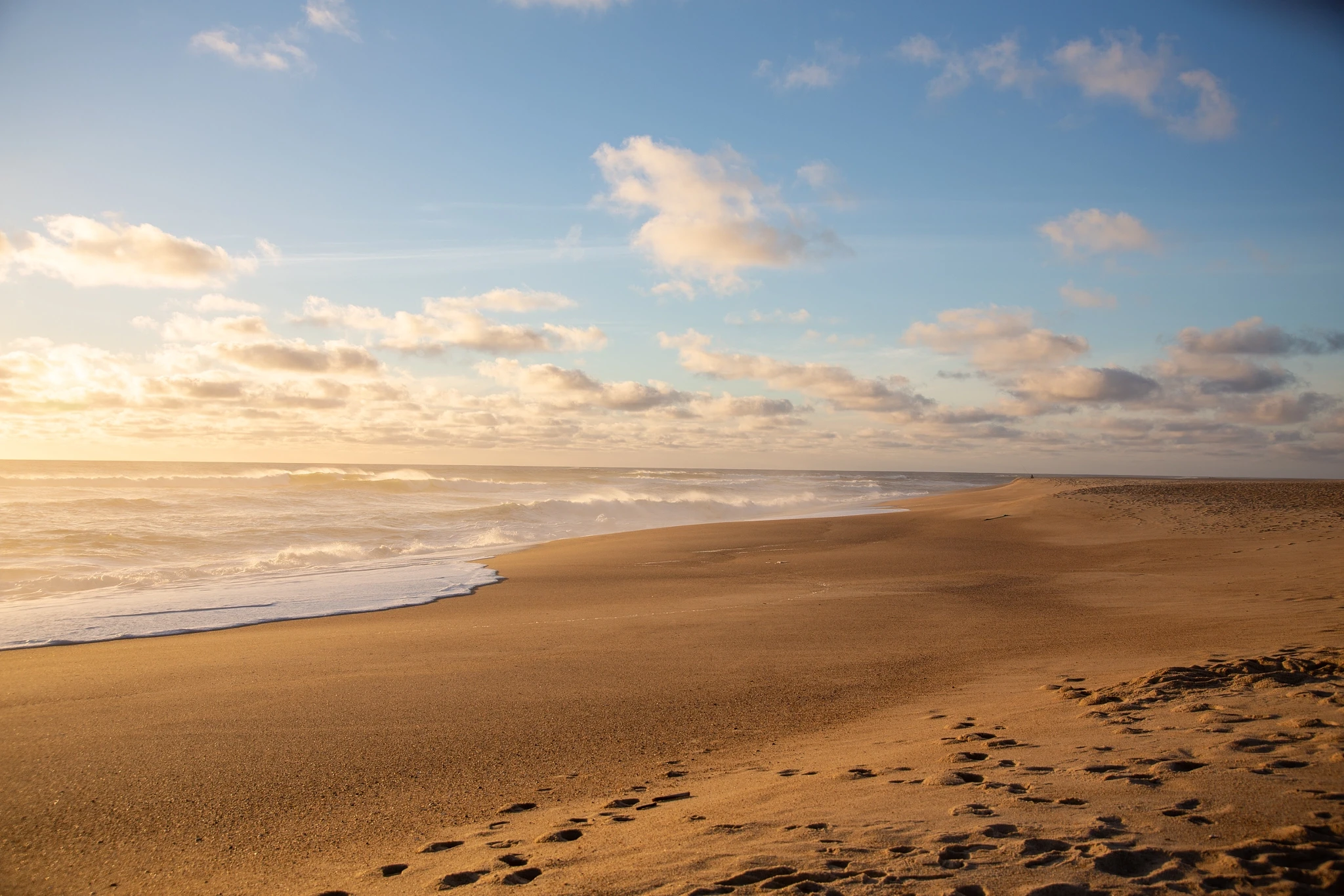 A vast empty beach catches the first warm light of day, gentle surf washing in from the left and a single line of footprints crossing the sand toward the glow on the horizon.