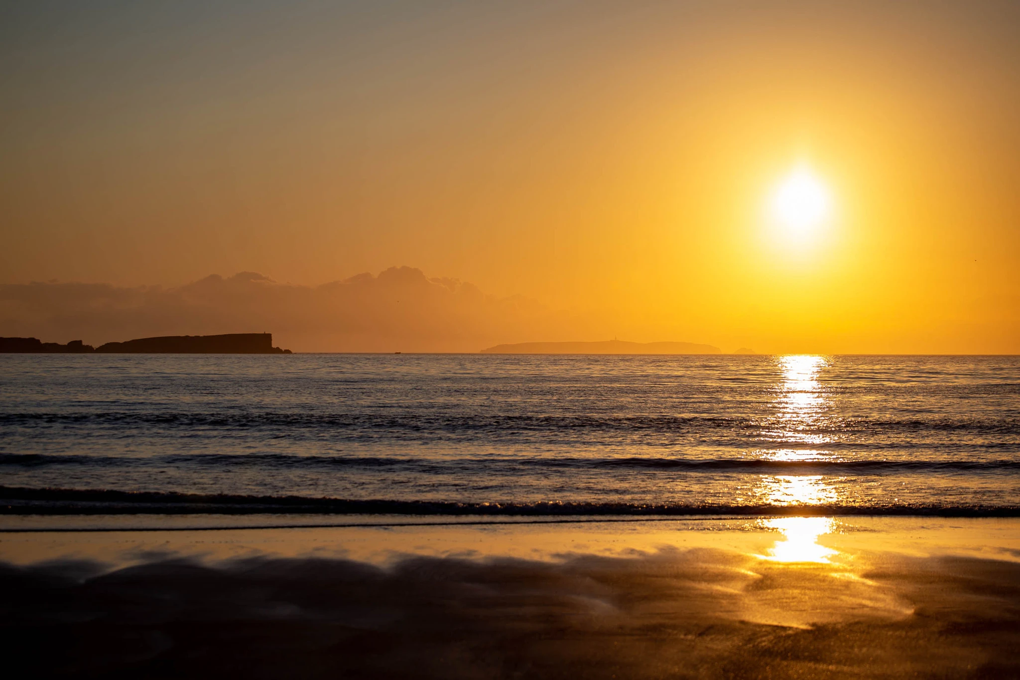 The setting sun casts a long path of gold across the flat ocean, a dark headland silhouetting on the left while the wet sand mirrors the light in warm amber pools.
