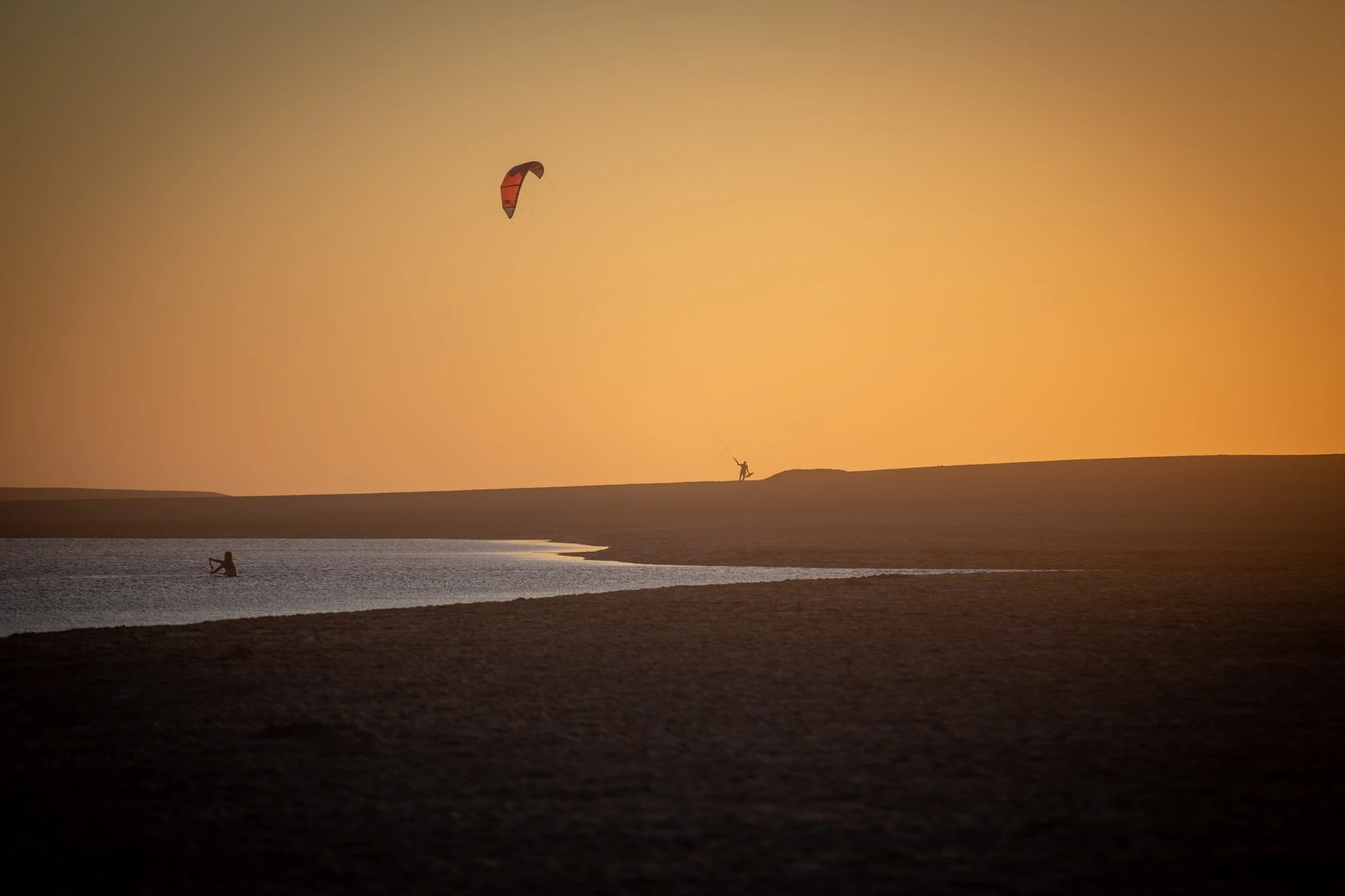 A single kitesurfer traces the surface of a shallow lagoon at deep orange sunset, their kite a dark crescent high in the sky, the flat landscape behind reduced to a thin silhouette.