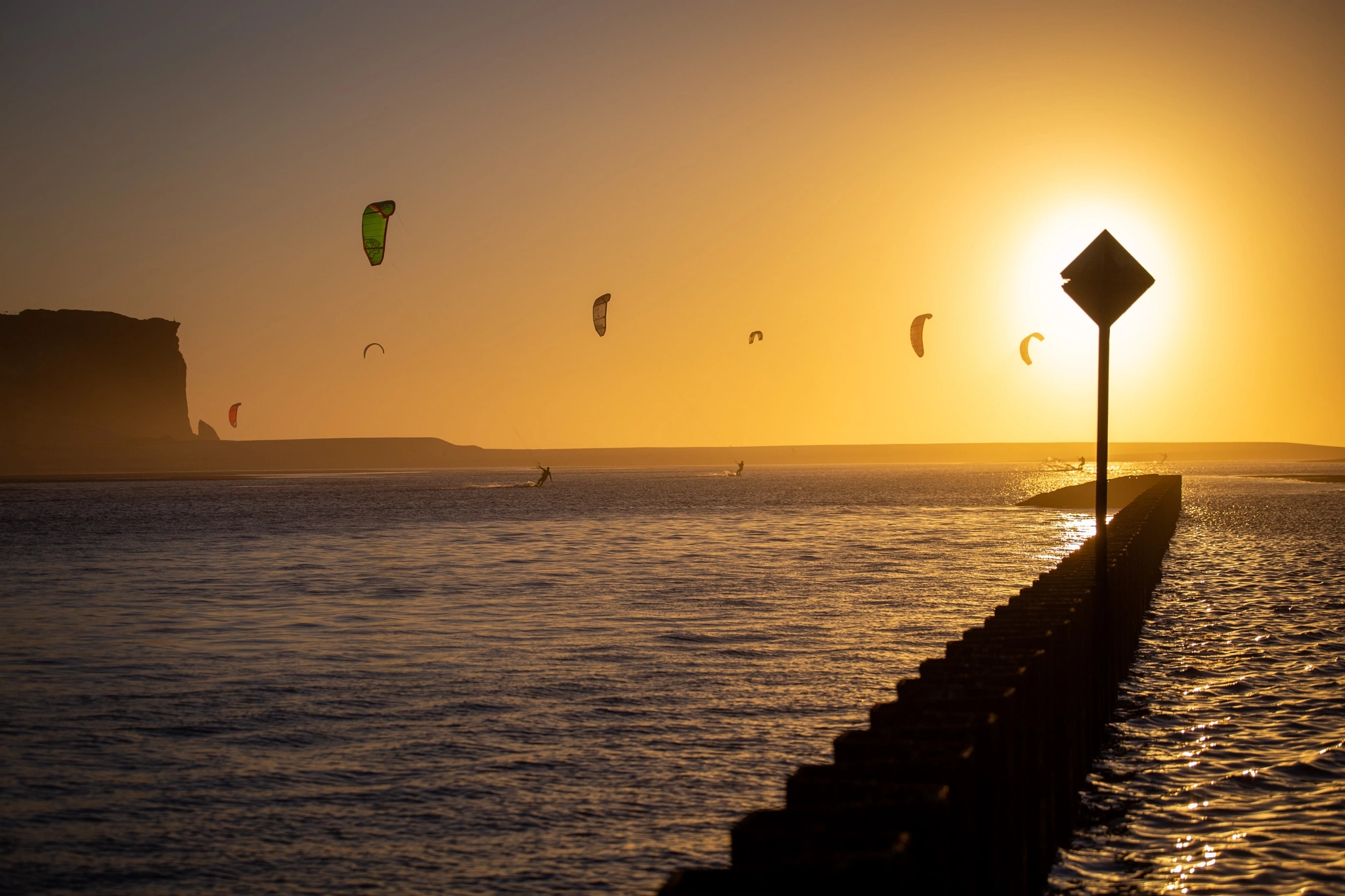 A sky crowded with kites hangs over a golden bay at sunset, the breakwater and its warning sign in silhouette while riders carve the water below in every direction.