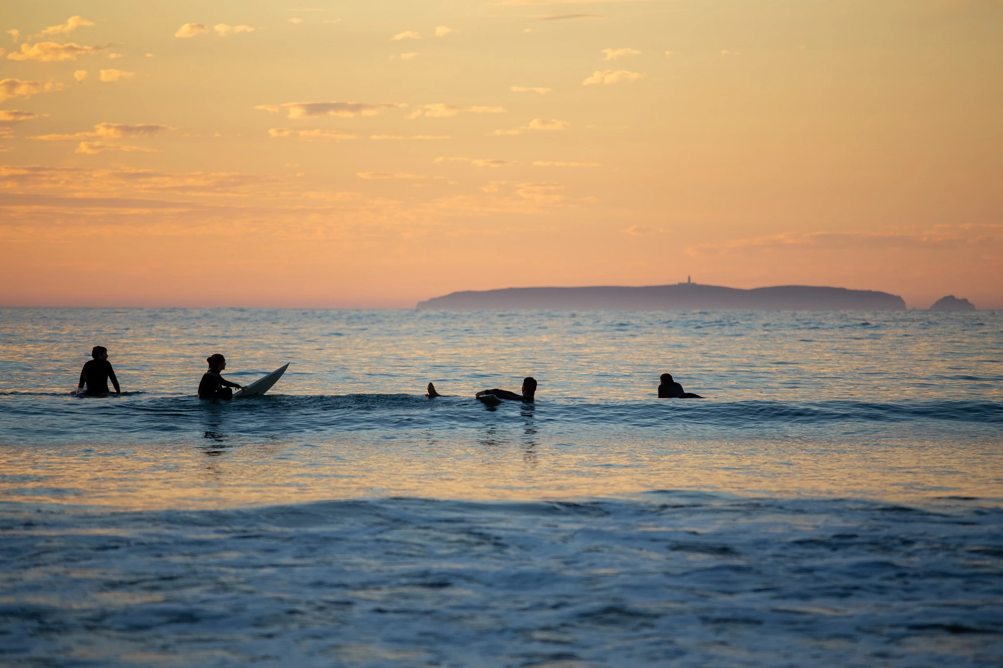 Four surfers lie on their boards in a flat calm lineup, the sun melting behind a distant island, the whole scene bathed in warm peach light with no waves in sight.