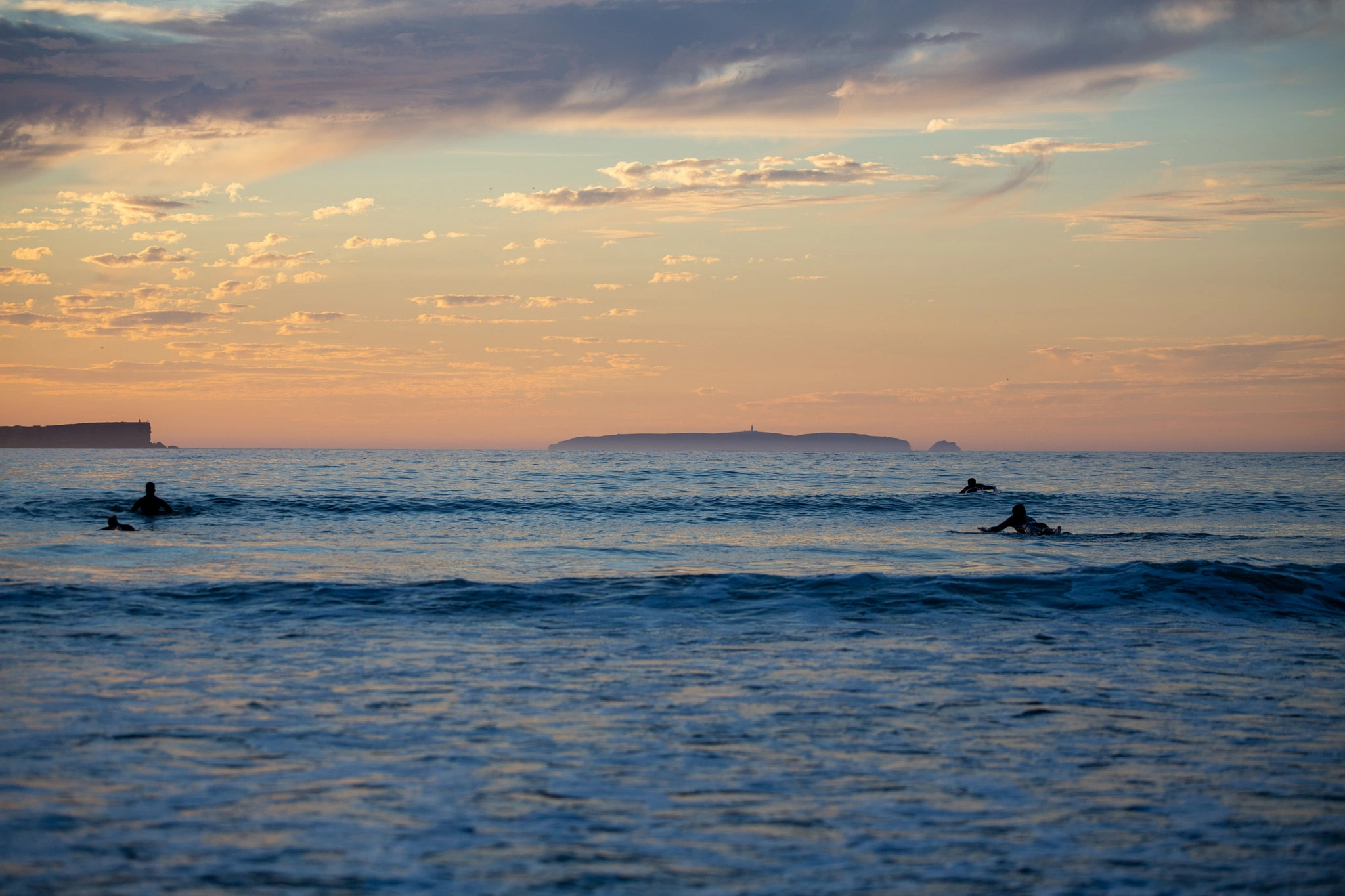Three surfers float silently at dusk, the sky a wash of peach and cobalt above a distant island, the Atlantic surface barely rippling as they wait.