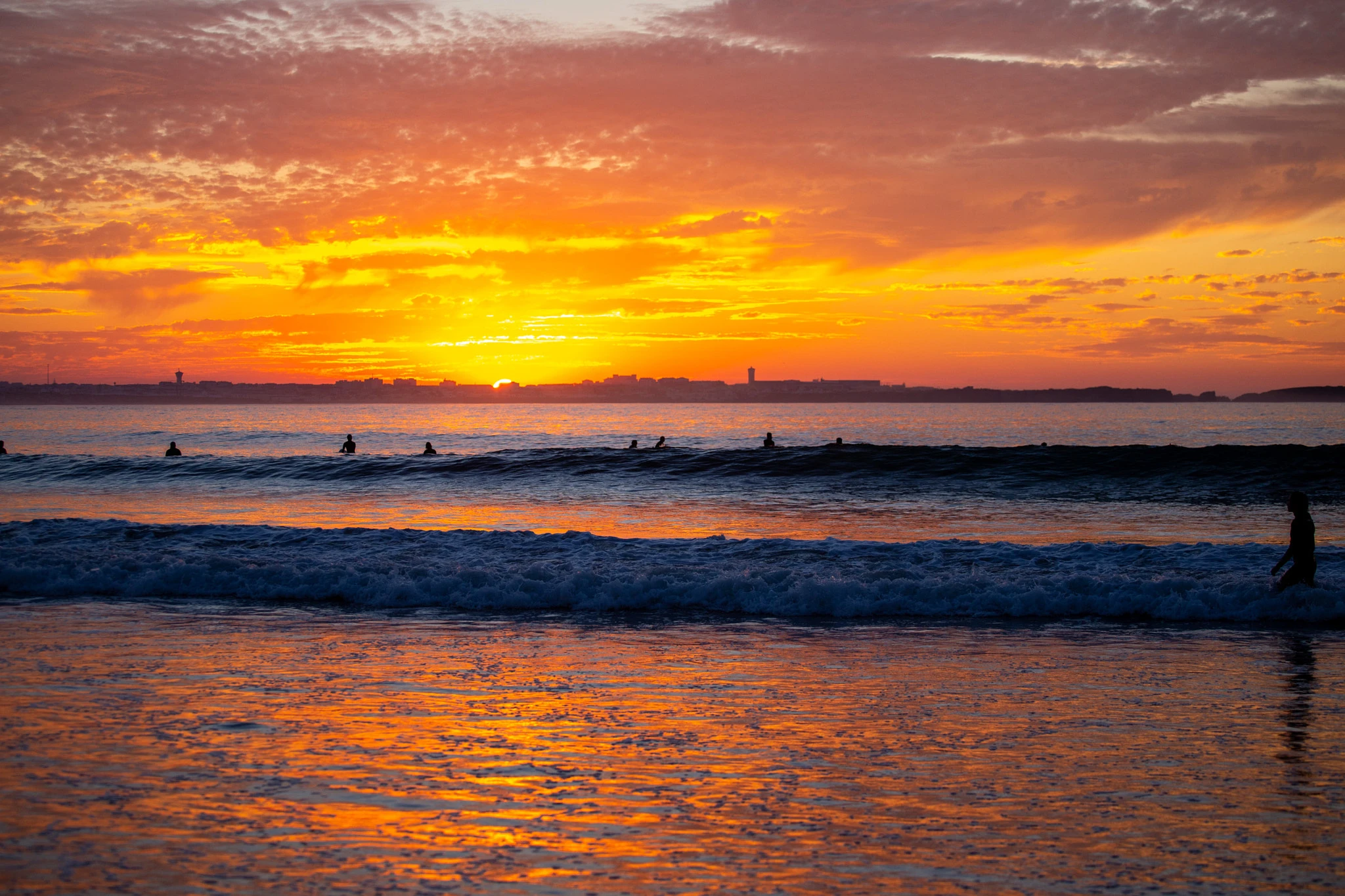 Surfers sit in the lineup as the sky blazes orange, the setting sun reflected in the wet sand at the shoreline where a lone figure wades in, the whole scene drenched in warm light.