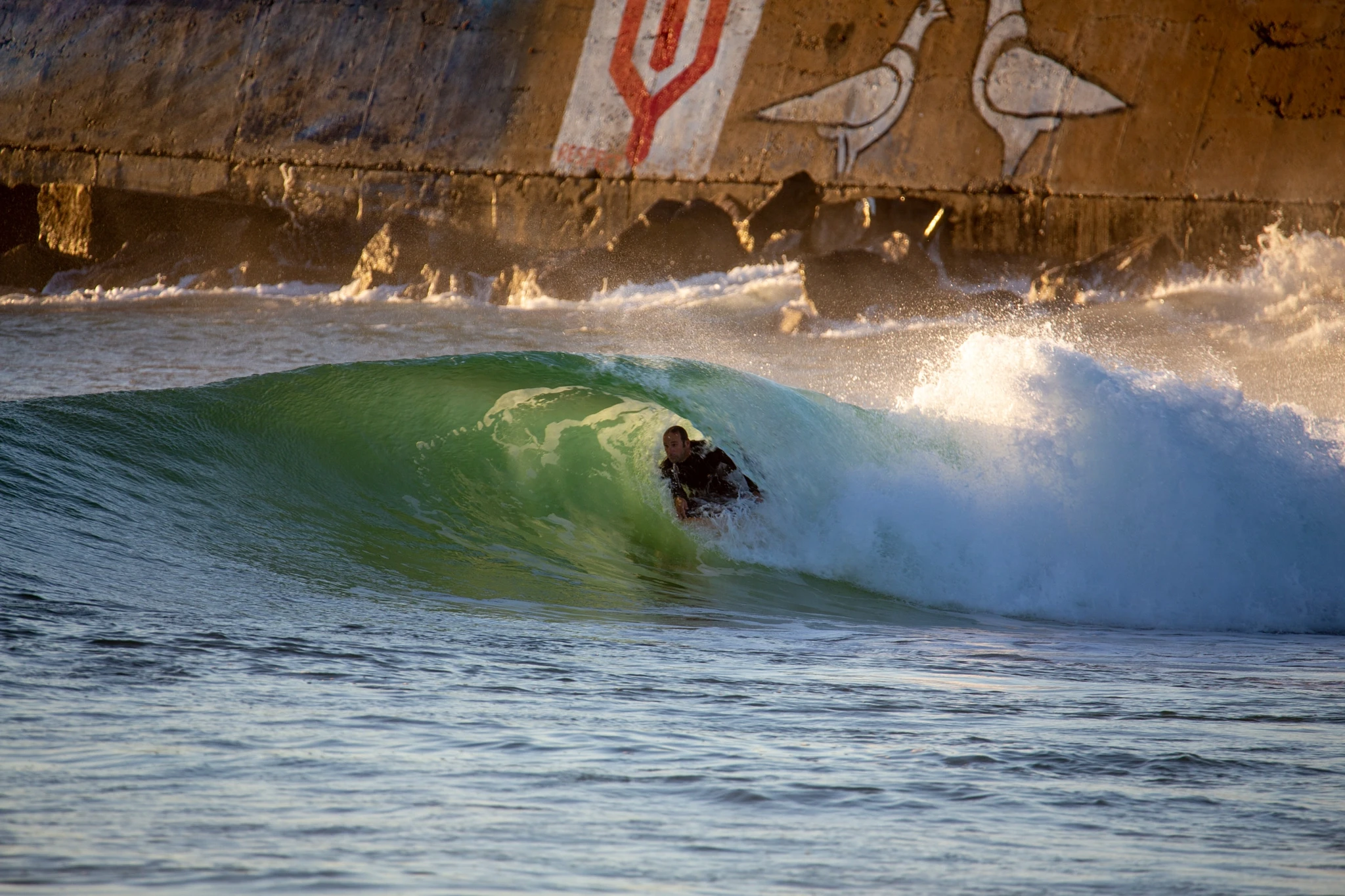 A bodyboarder is fully encased in a glassy emerald barrel, the wave lip overhead, a graffiti-painted sea wall visible behind the surging white foam — textbook tube riding.