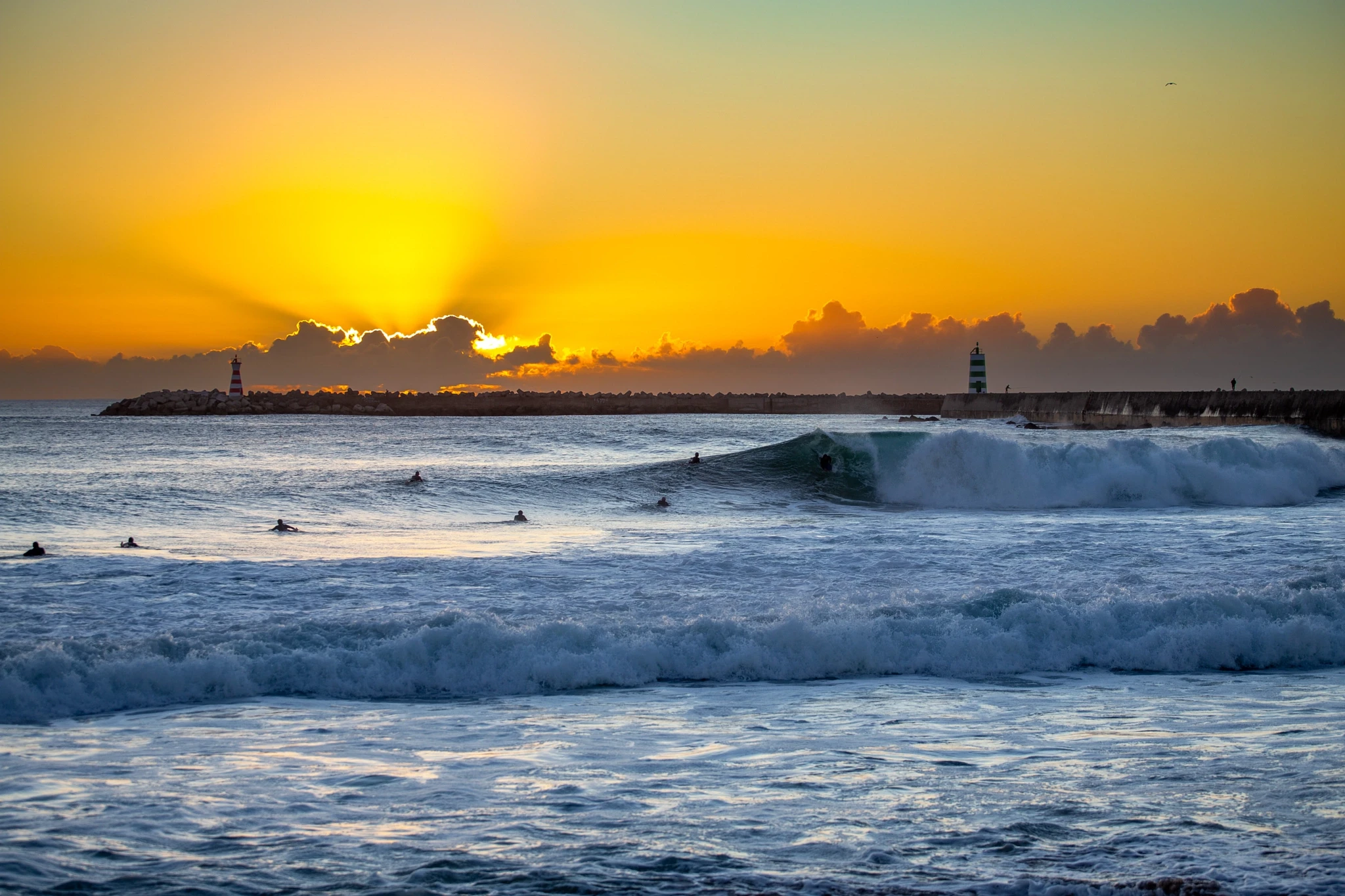A surfer catches a wave as the sky turns to gold, two lighthouse towers standing sentinel on the breakwater behind while other surfers drift in the glowing water.