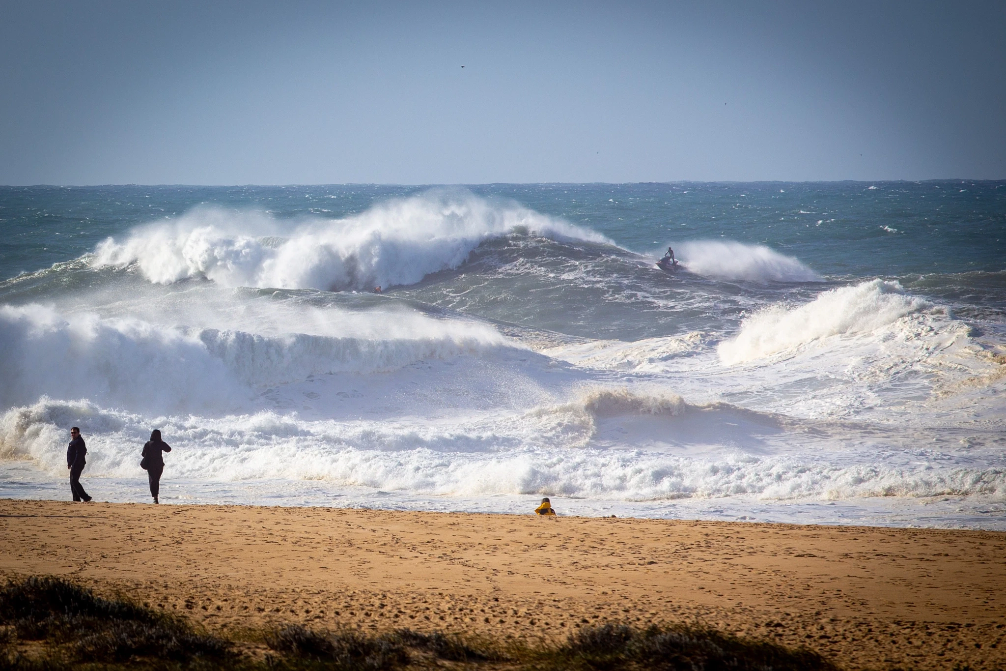 Two figures stand at the shoreline watching monstrous storm waves detonate on the beach, a jet ski buried in the white water behind them — the raw power of the Atlantic on full display.