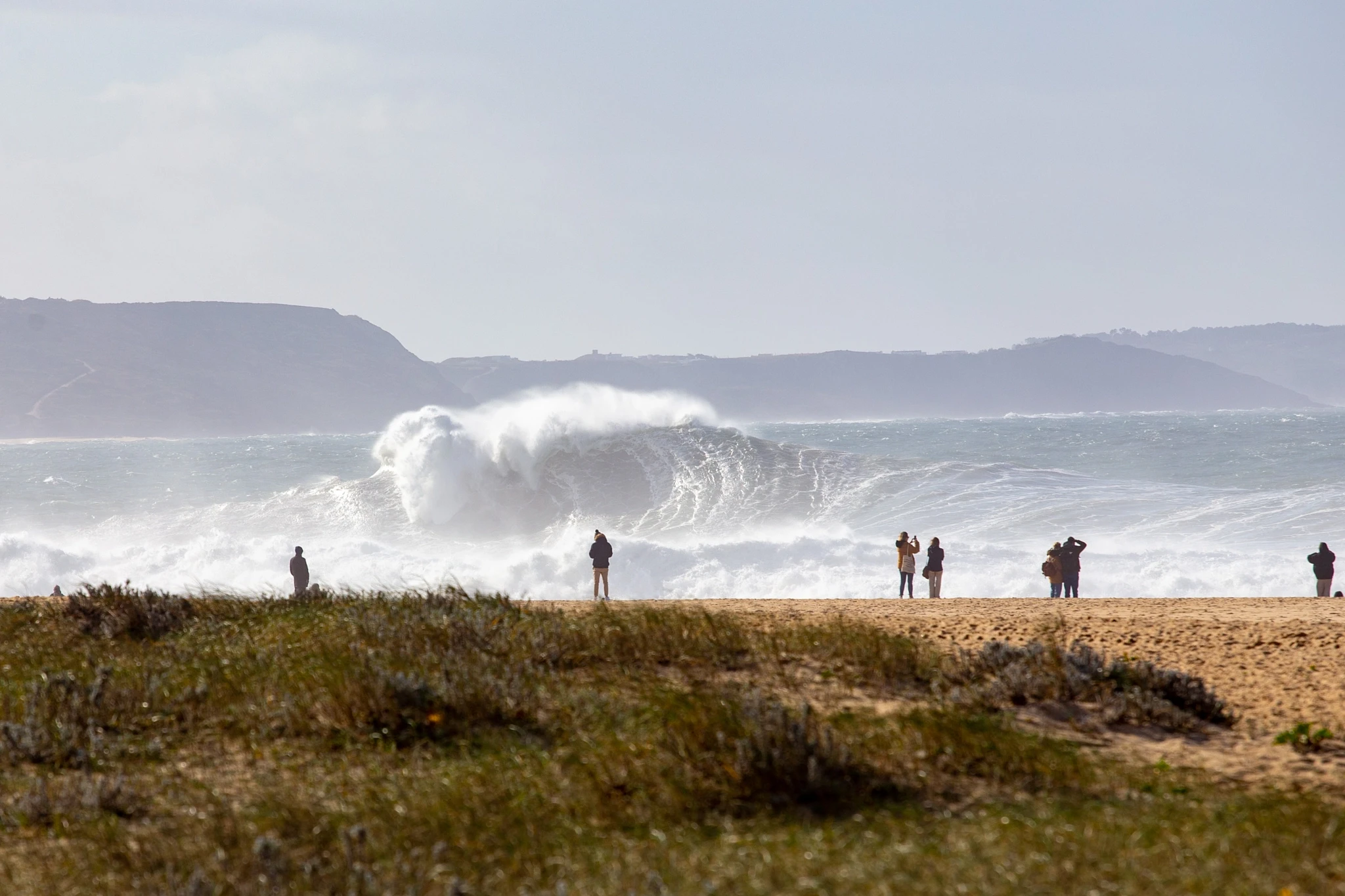 A small group of spectators watches from the dunes as an enormous wave detonates in the shallows, spray thrown high into the air, dark mountains lining the horizon behind.