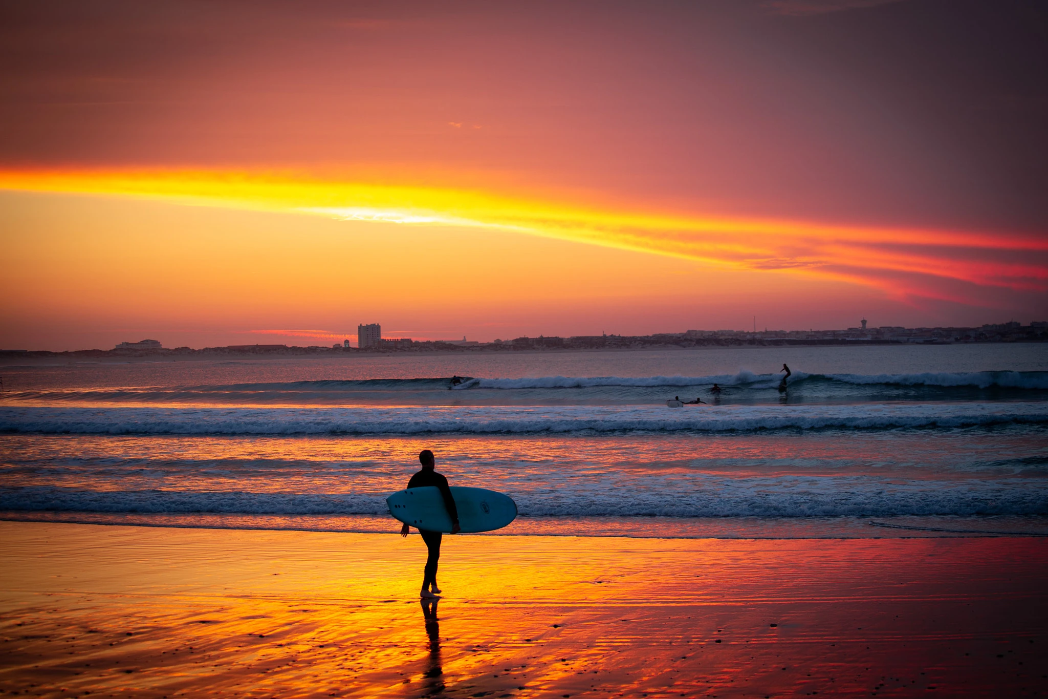 A surfer heads across the wet sand at the end of the day, board under arm, their figure silhouetted against a blazing sky of red, yellow and orange, the city skyline a dark band behind.