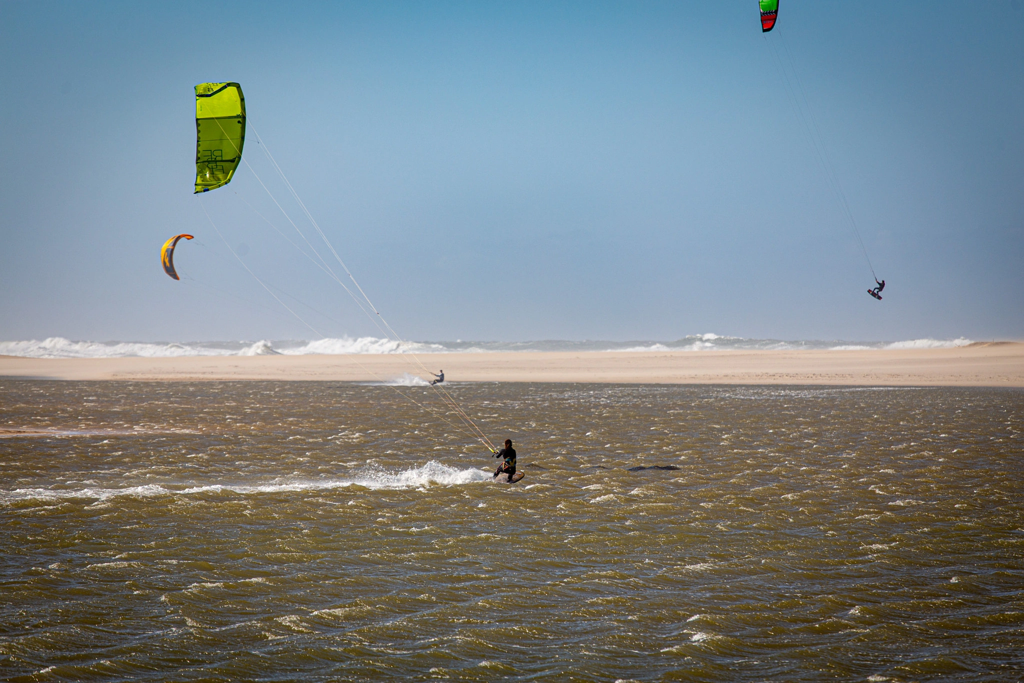 Three kitesurfers work the rough, wind-whipped water during a blow, their canopies taut against the effort, a long sandy beach stretching behind under a flat grey-blue sky.