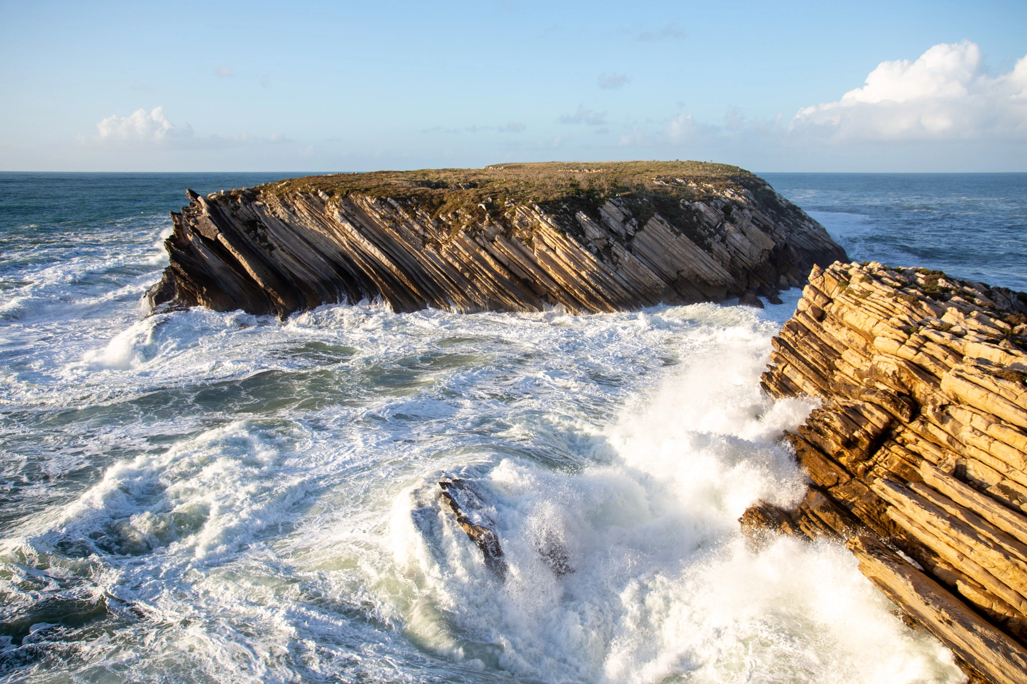 The steeply angled strata of coastal rocks meet the sea in a cascade of white water, a flat-topped island rising beyond and a bright sky with racing clouds overhead.