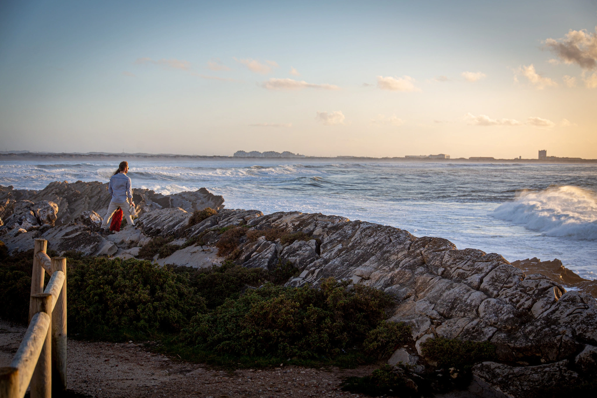 A solitary figure picks their way across a rocky coastal headland to watch rough surf rolling in, the warm evening light catching the water while a town glows softly on the distant shore.
