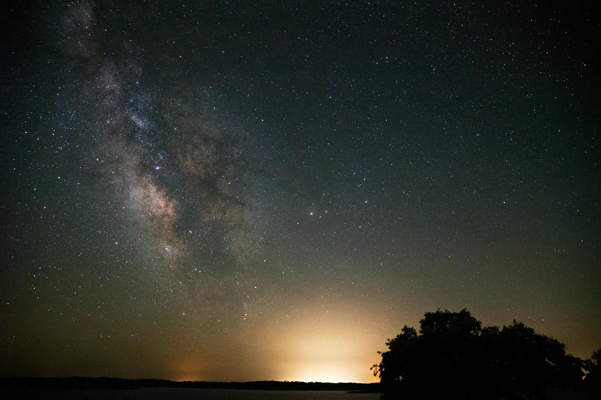 The dense core of the Milky Way arches across the sky above a darkened landscape, tree silhouettes on the right and a distant city glow warming the horizon in amber.