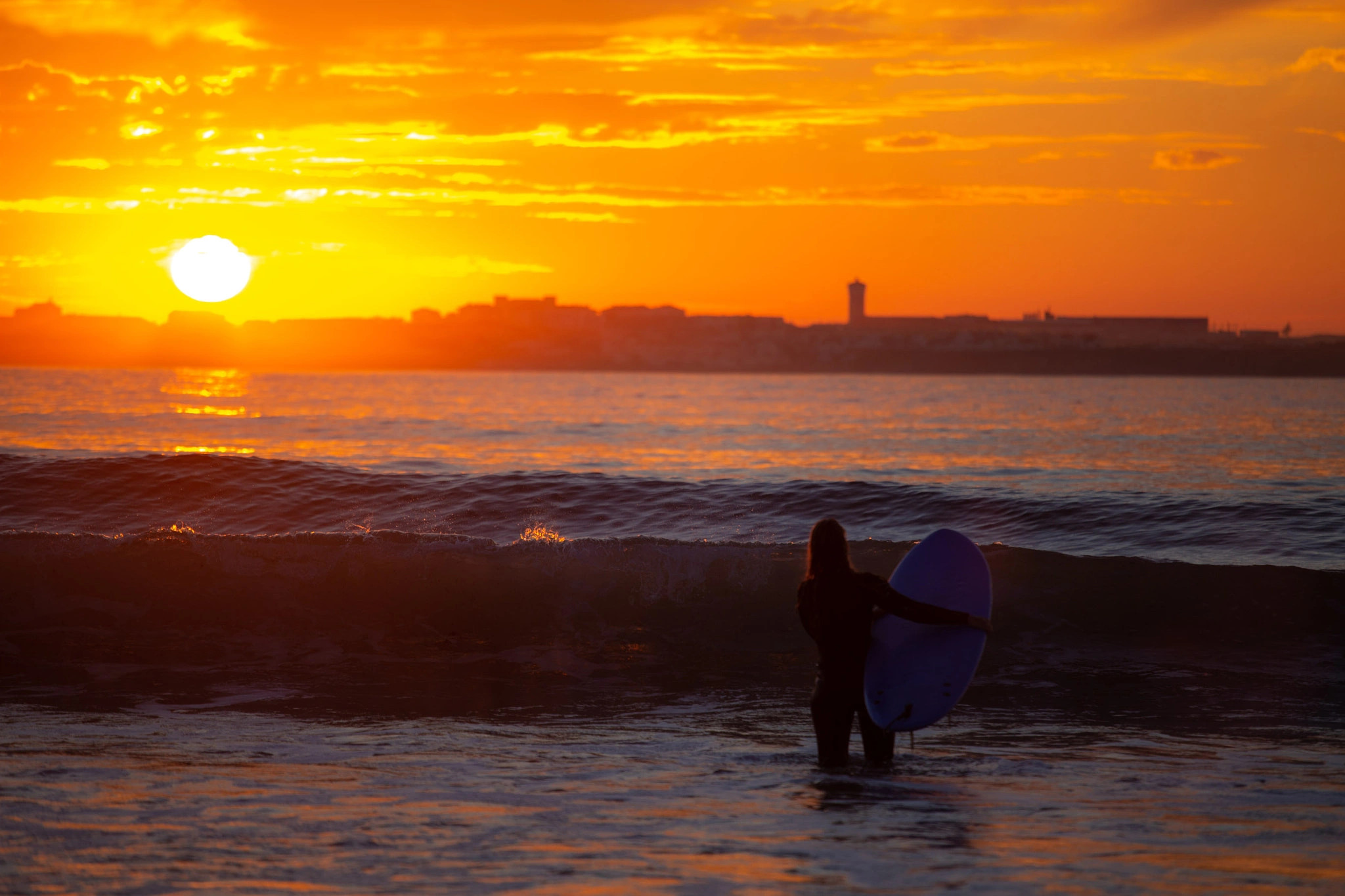 A female surfer stands waist-deep in the shallows, board pressed to her side, the sky behind a wall of orange fire, a coastal town silhouetted across the bay.