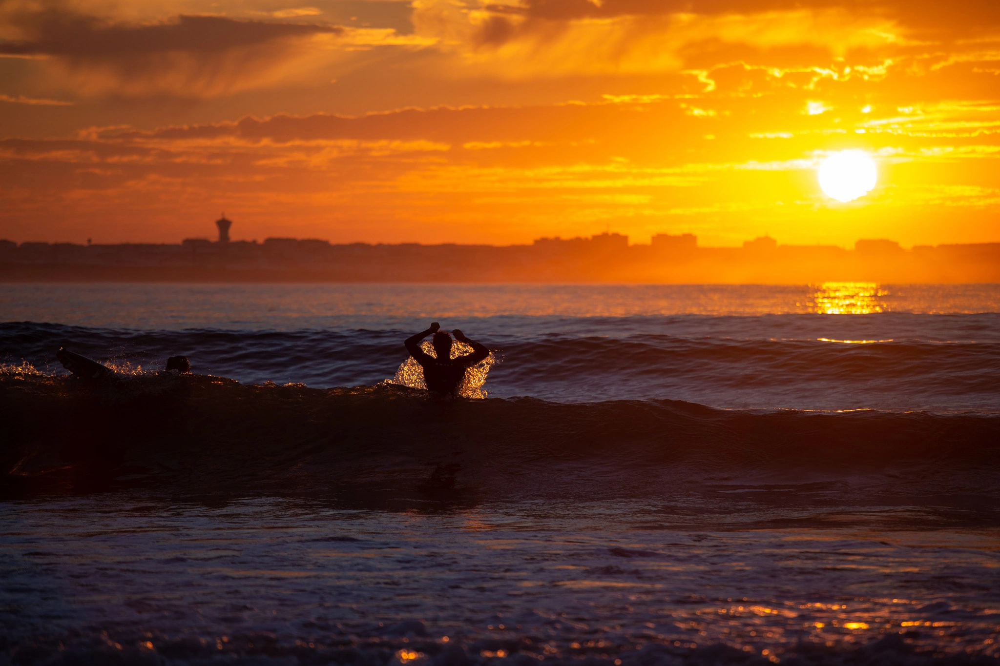 A surfer is swallowed by a breaking wave at the moment the sun flares full on the horizon, the city skyline dark behind, the sky alive with orange and gold.