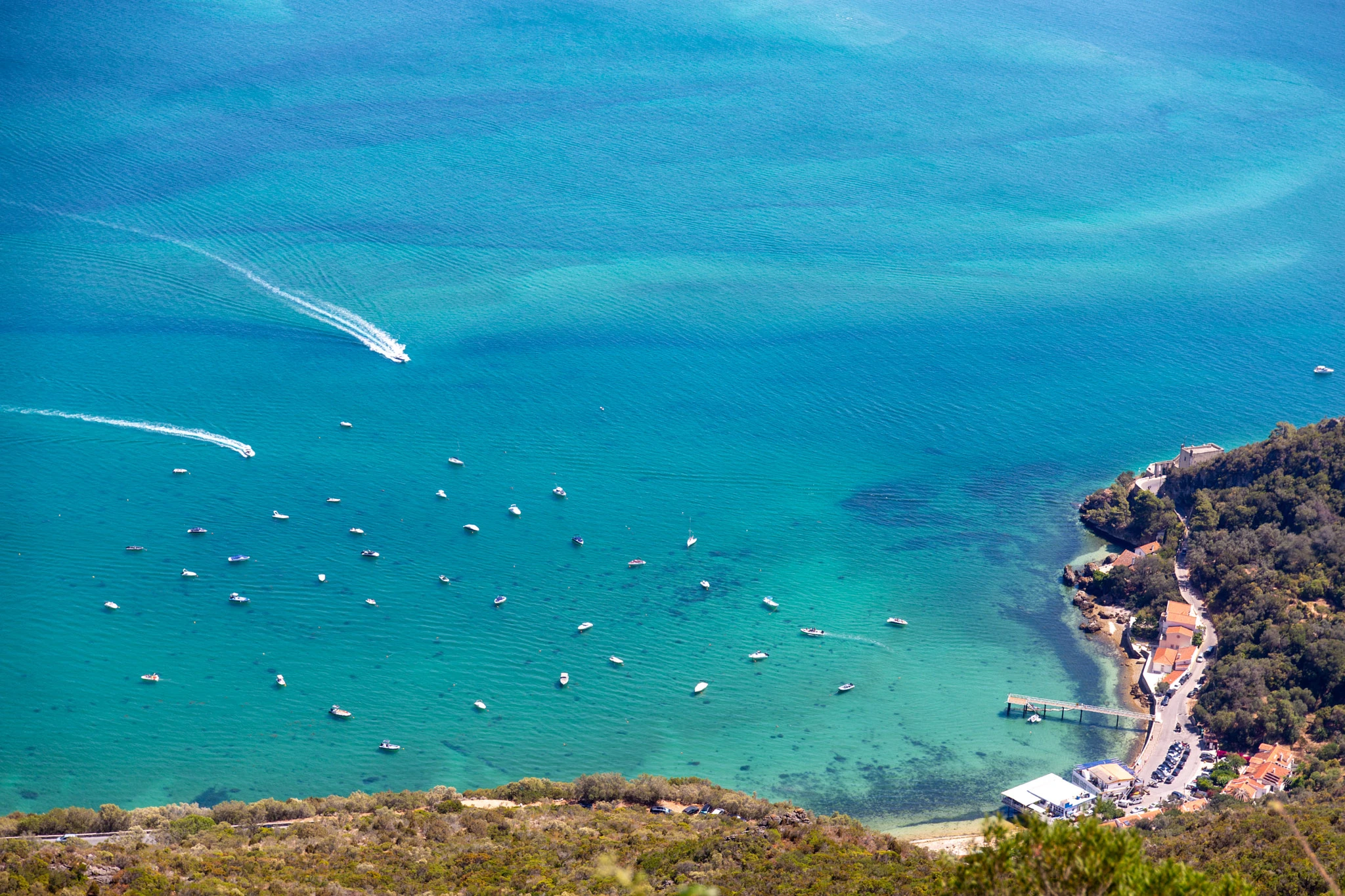 Seen from high on the cliffs above, a dozen small boats lie anchored in an impossibly turquoise bay, a speedboat's wake cutting through the shallows where a tiny village clings to the shore.