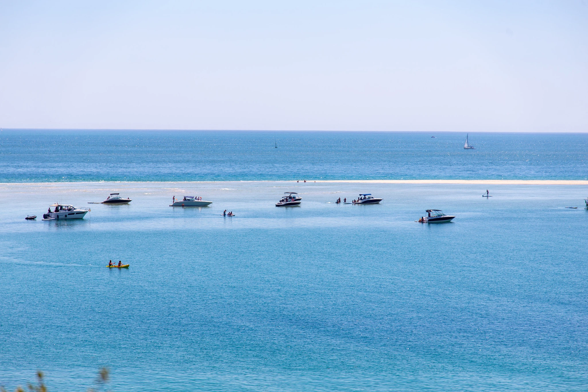 Several powerboats cluster around a submerged sandbar in a glittering turquoise bay, a yellow kayak in the foreground and two sailboats drifting on the hazy horizon beyond.