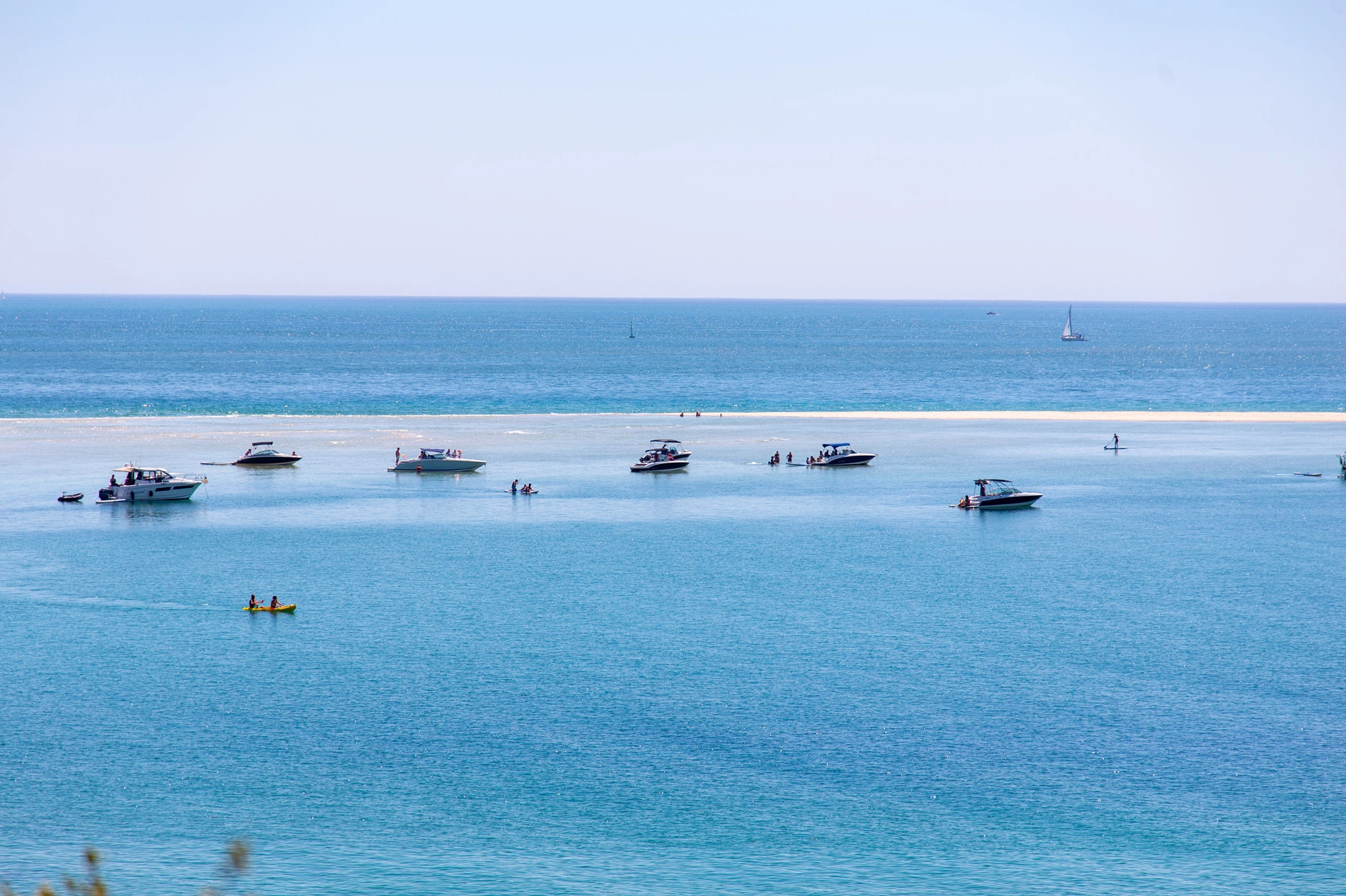 Several powerboats cluster around a submerged sandbar in a glittering turquoise bay, a yellow kayak in the foreground and two sailboats drifting on the hazy horizon beyond.