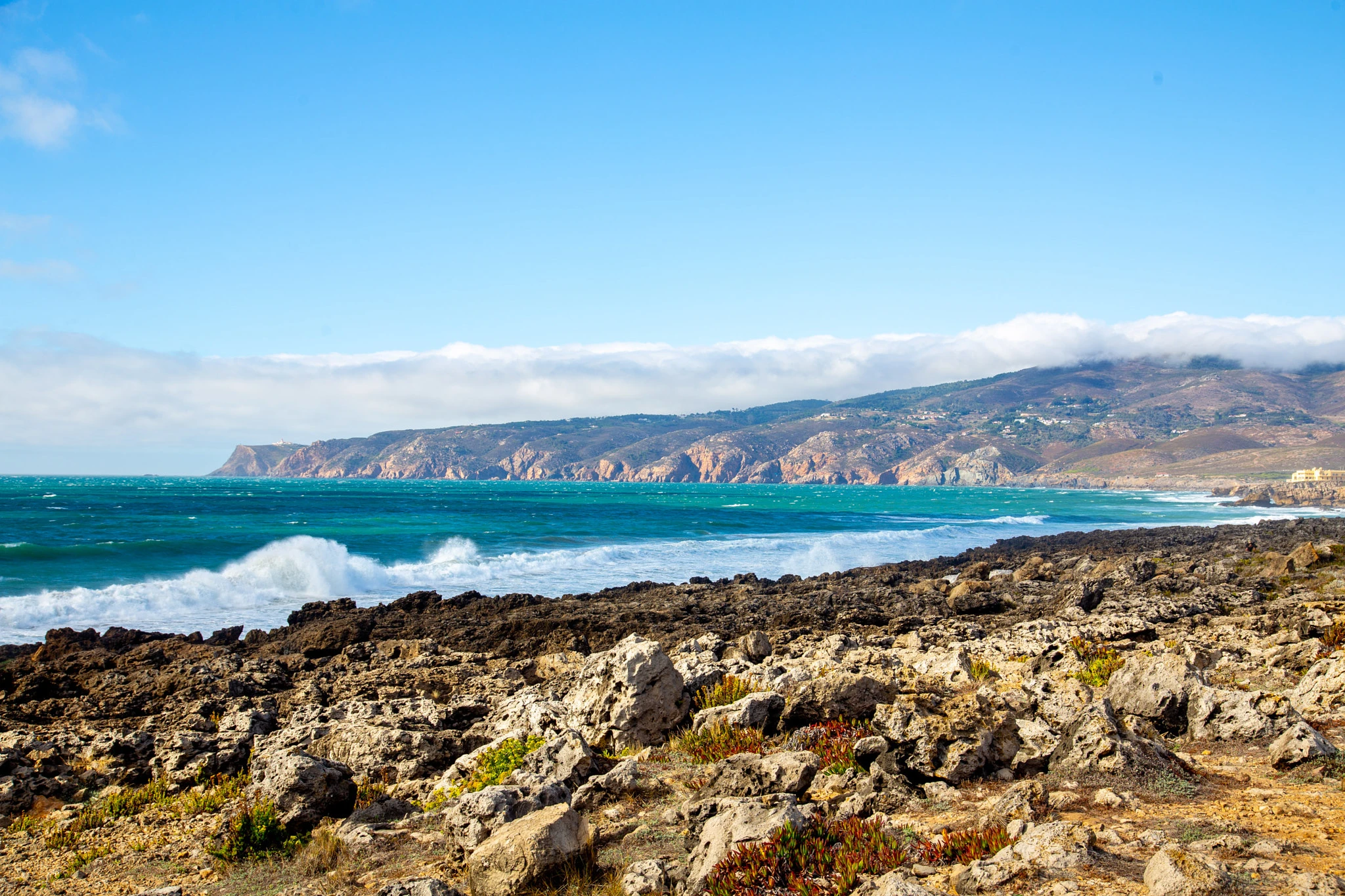 Vivid turquoise waves break over a rugged rocky foreground, the dramatic cliff line of the Sintra coast stretching far into the distance under a layer of low cloud.