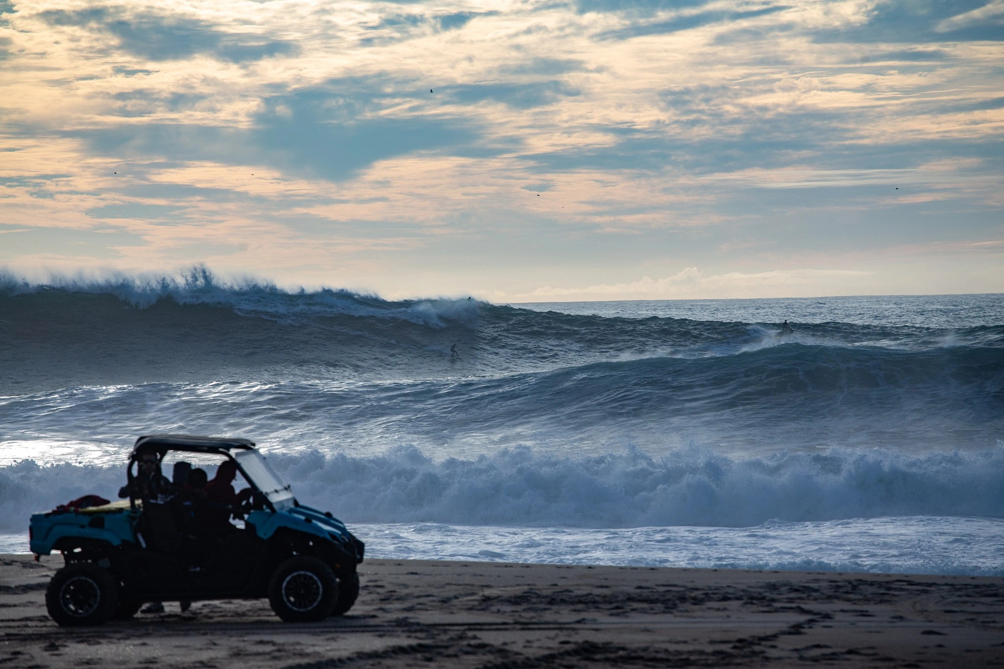 A beach patrol buggy is parked on the sand watching a surfer ride enormous waves — walls of white water that dwarf everything, the sky heavy and overcast.