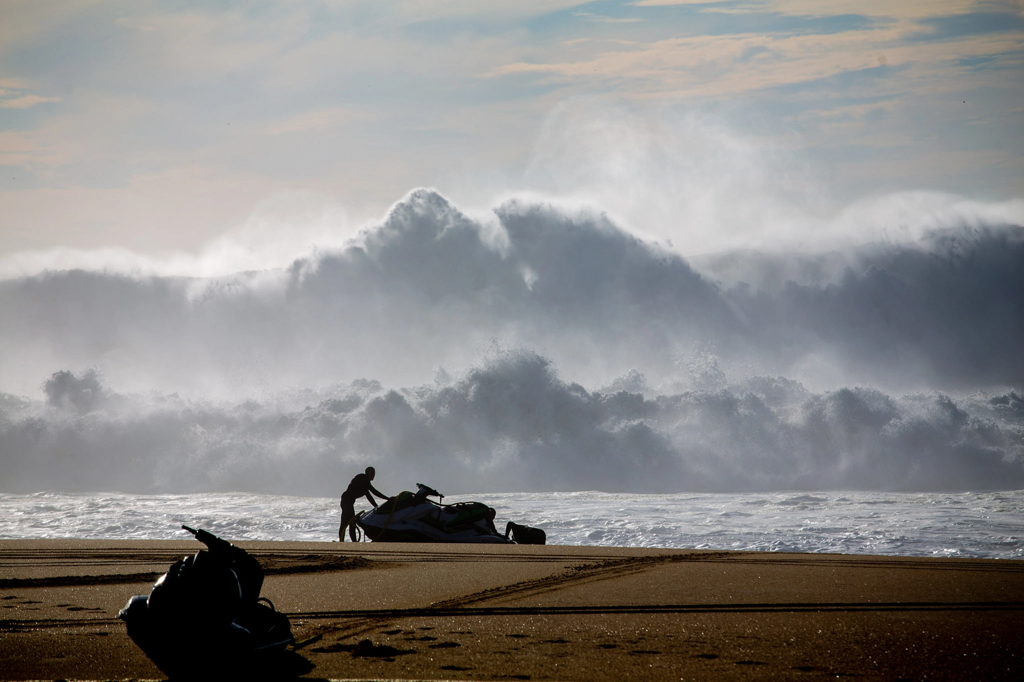 A figure works with a jet ski at the waterline as enormous waves detonate behind them, mist and spray filling the air in a towering white wall of force.