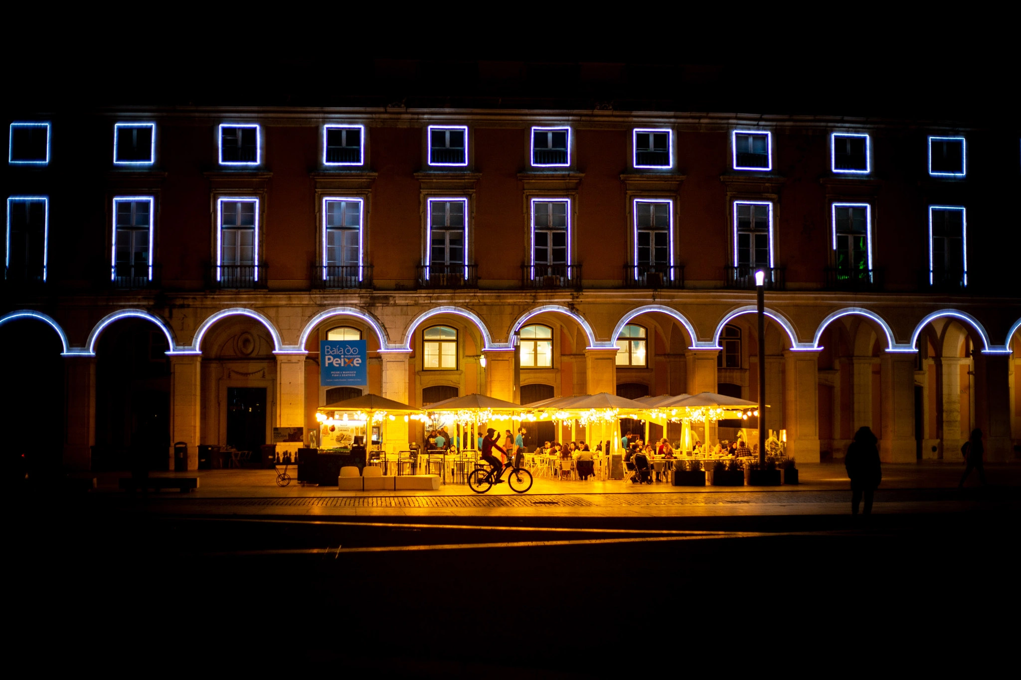 The grand arched façade of Praça do Comércio glows with warm restaurant light and blue LED outlines, a cyclist crossing in front — Lisbon at its most theatrical.