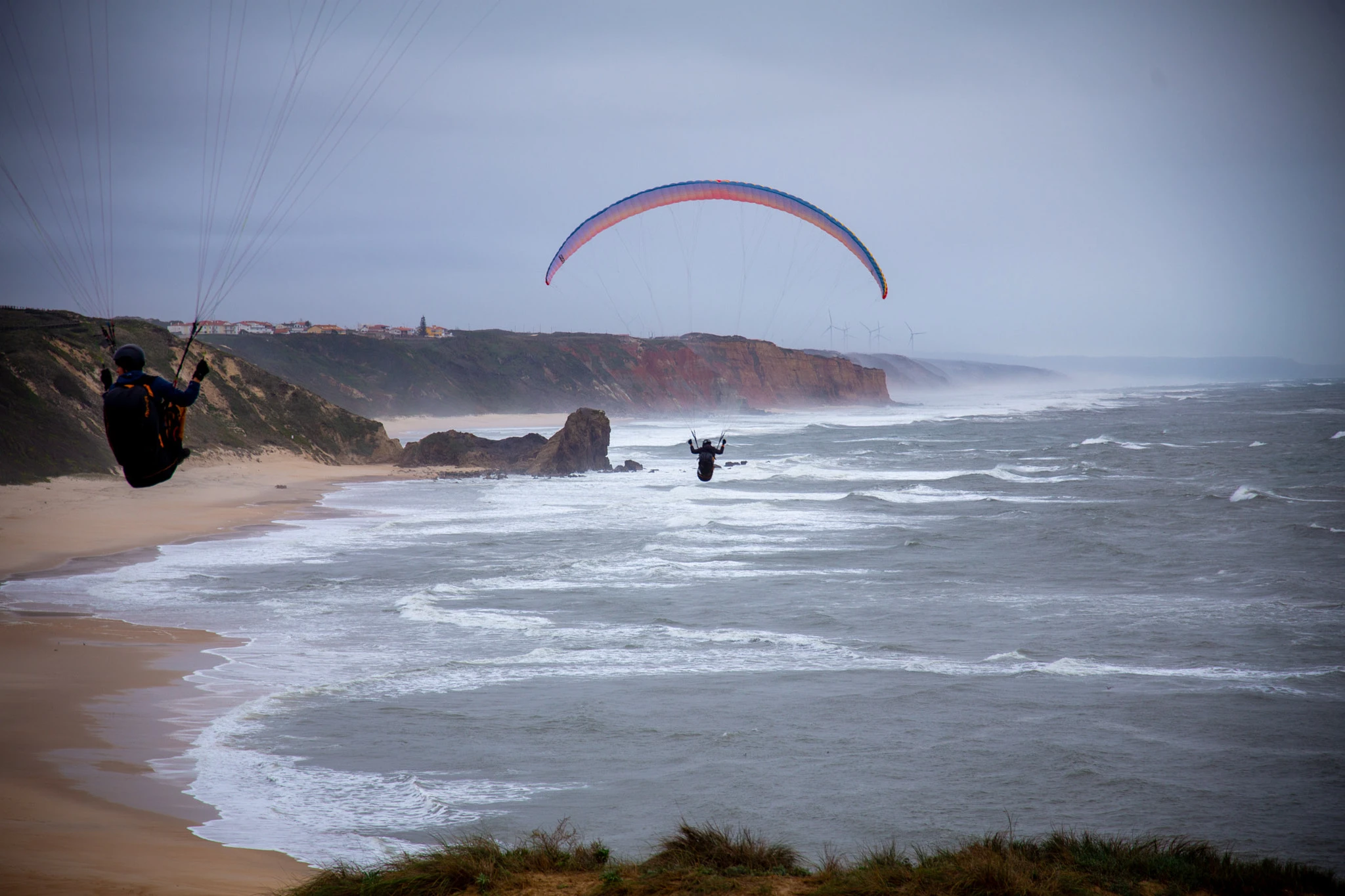 Two paragliders soar low over the beach, the red sandstone cliffs of the Portuguese coast stretching behind them, wind turbines on the distant hills, the Atlantic grey and rough below.