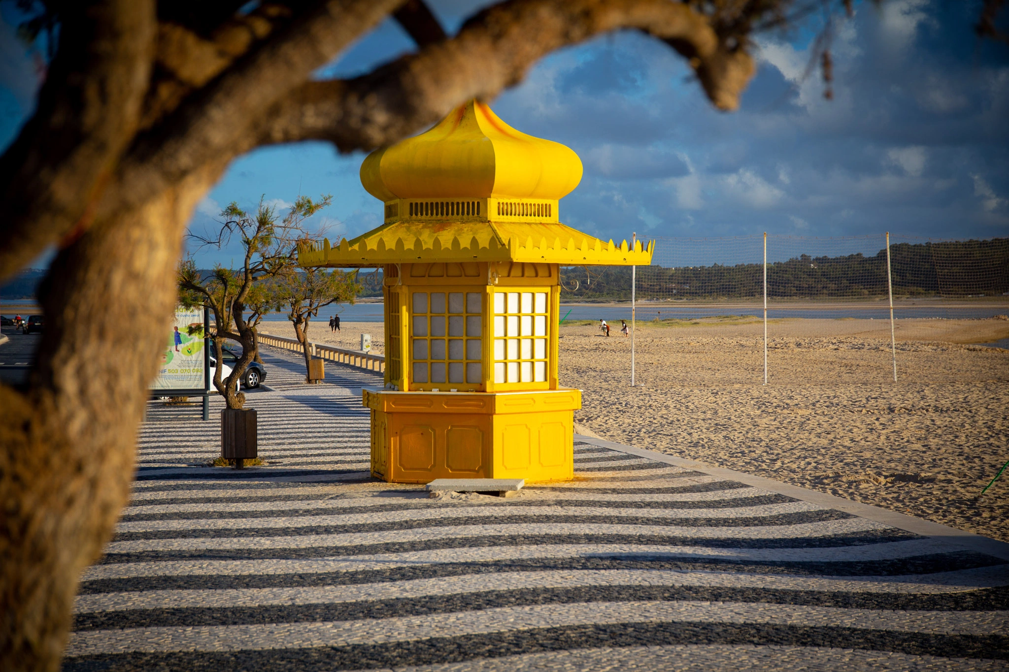 A vivid yellow oriental-style kiosk stands on a traditional Portuguese mosaic promenade, the beach stretching beyond and a twisted tree branch framing the scene from above.