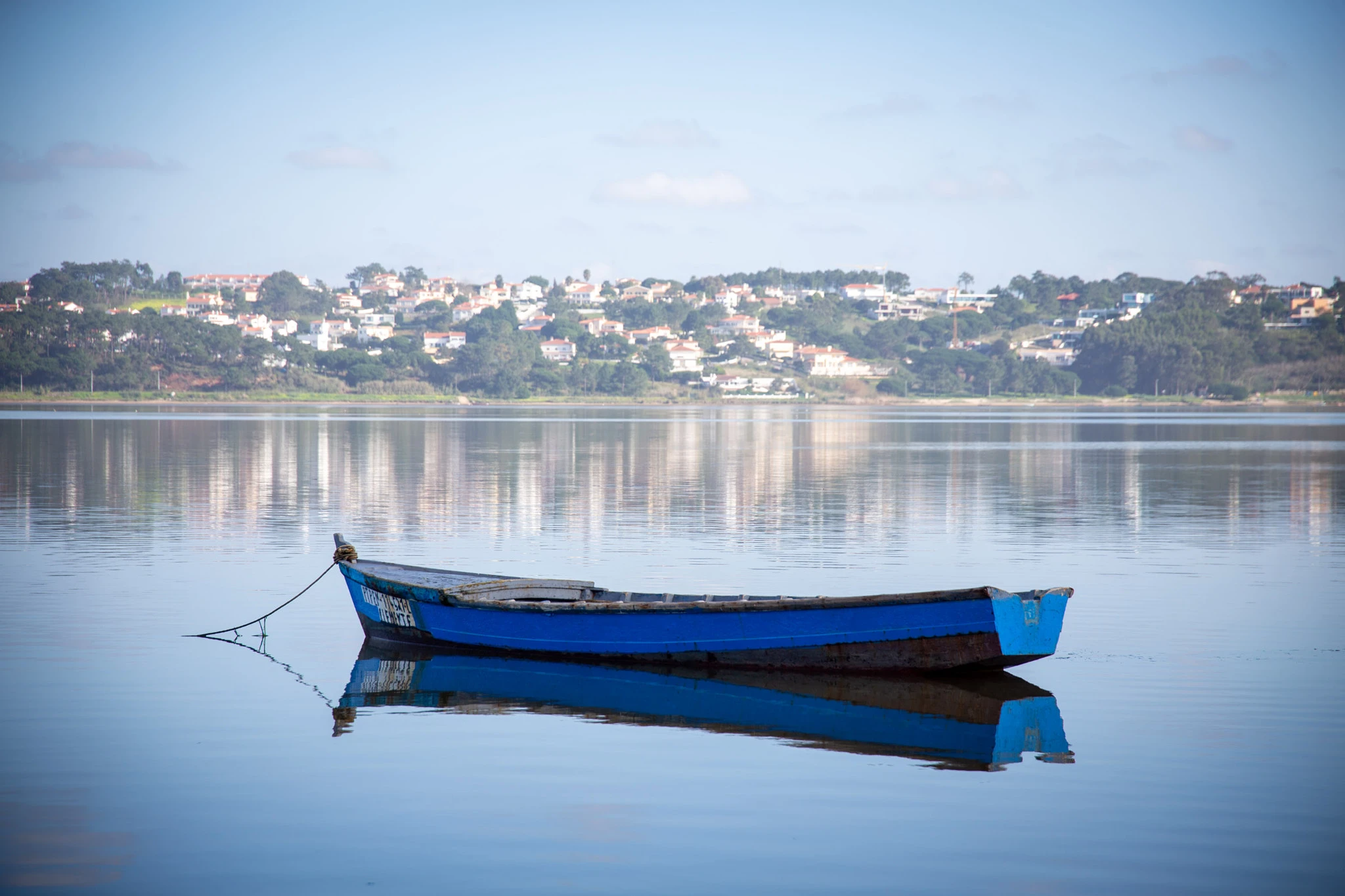 A worn blue rowing boat is moored on a lagoon so still it creates a perfect mirror image, a white-washed Portuguese village nestled in green trees on the far bank.