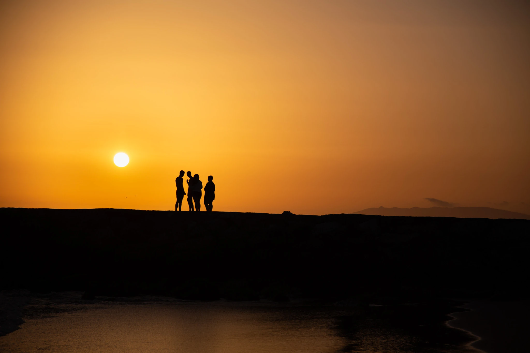 Three people stand at the edge of a flat dark headland as the sun sets in a clean blaze of orange, a low mountain range tracing the horizon and the ocean below catching the last light.