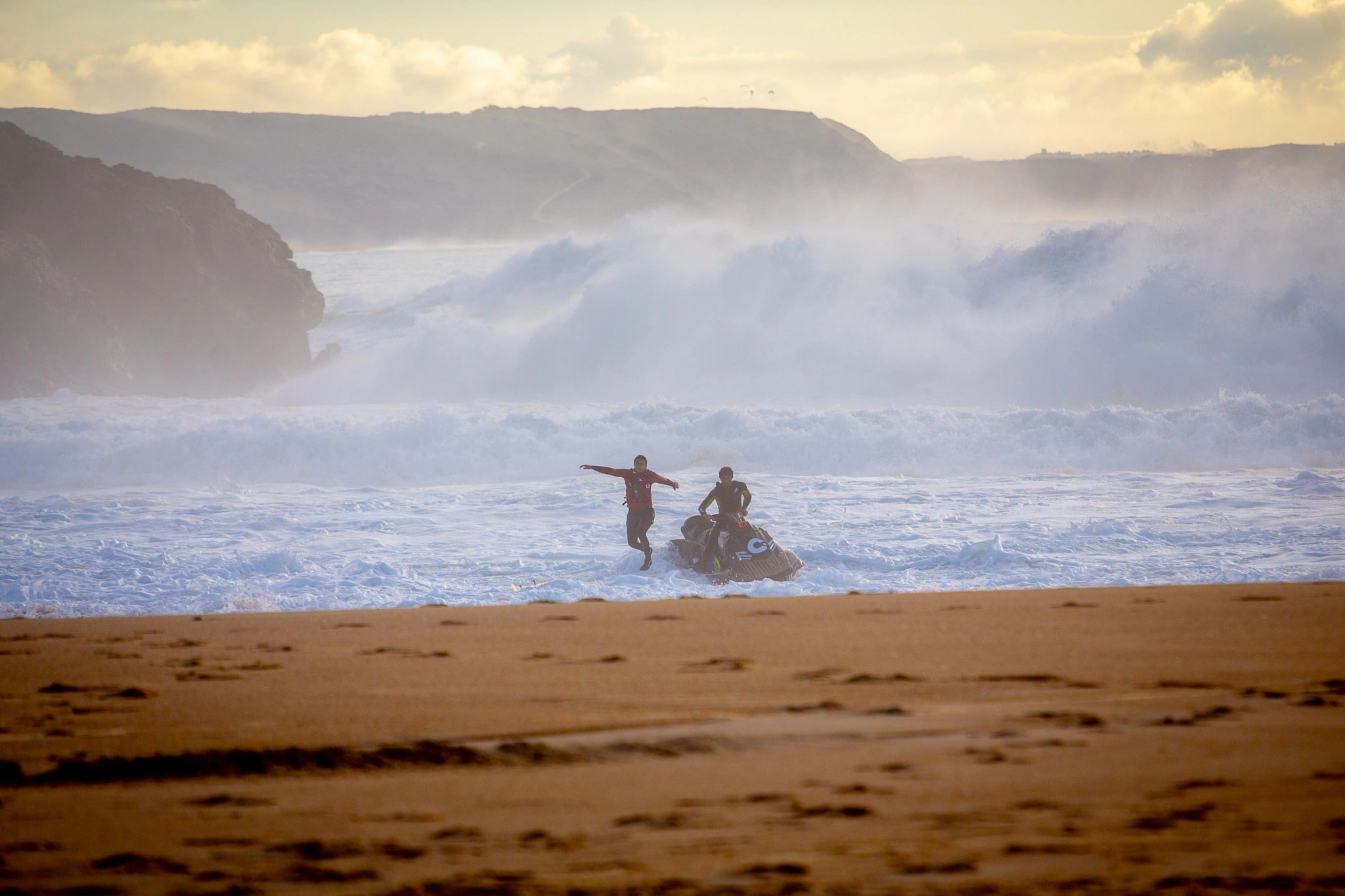 Two figures stand by a jet ski at the water's edge, facing massive waves that crash against the base of the cliffs beyond — a moment of calm focus before launching into chaos.