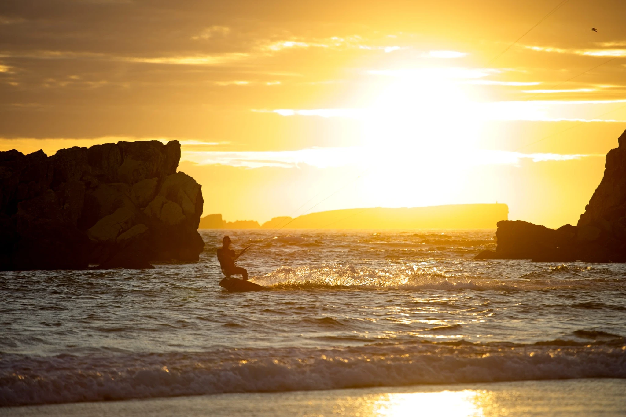 A kitesurfer skims through the channel between two dark sea stacks at blazing golden hour, an island glowing on the horizon beyond — framed perfectly by the natural rock gate.