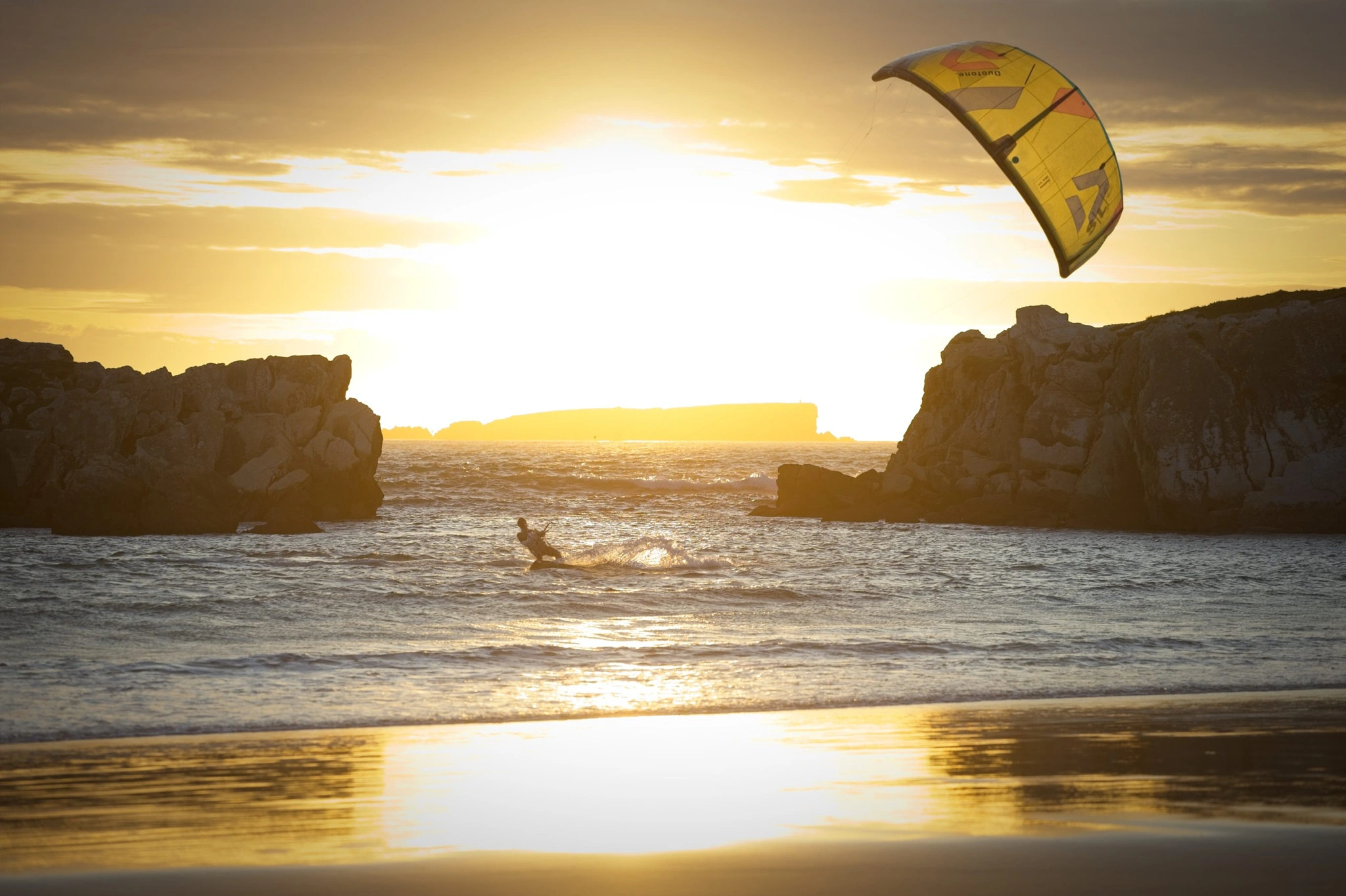 A kitesurfer with a vivid yellow kite carves through the golden sea between two rock formations at sunset, the wet sand perfectly reflecting the blazing sky.